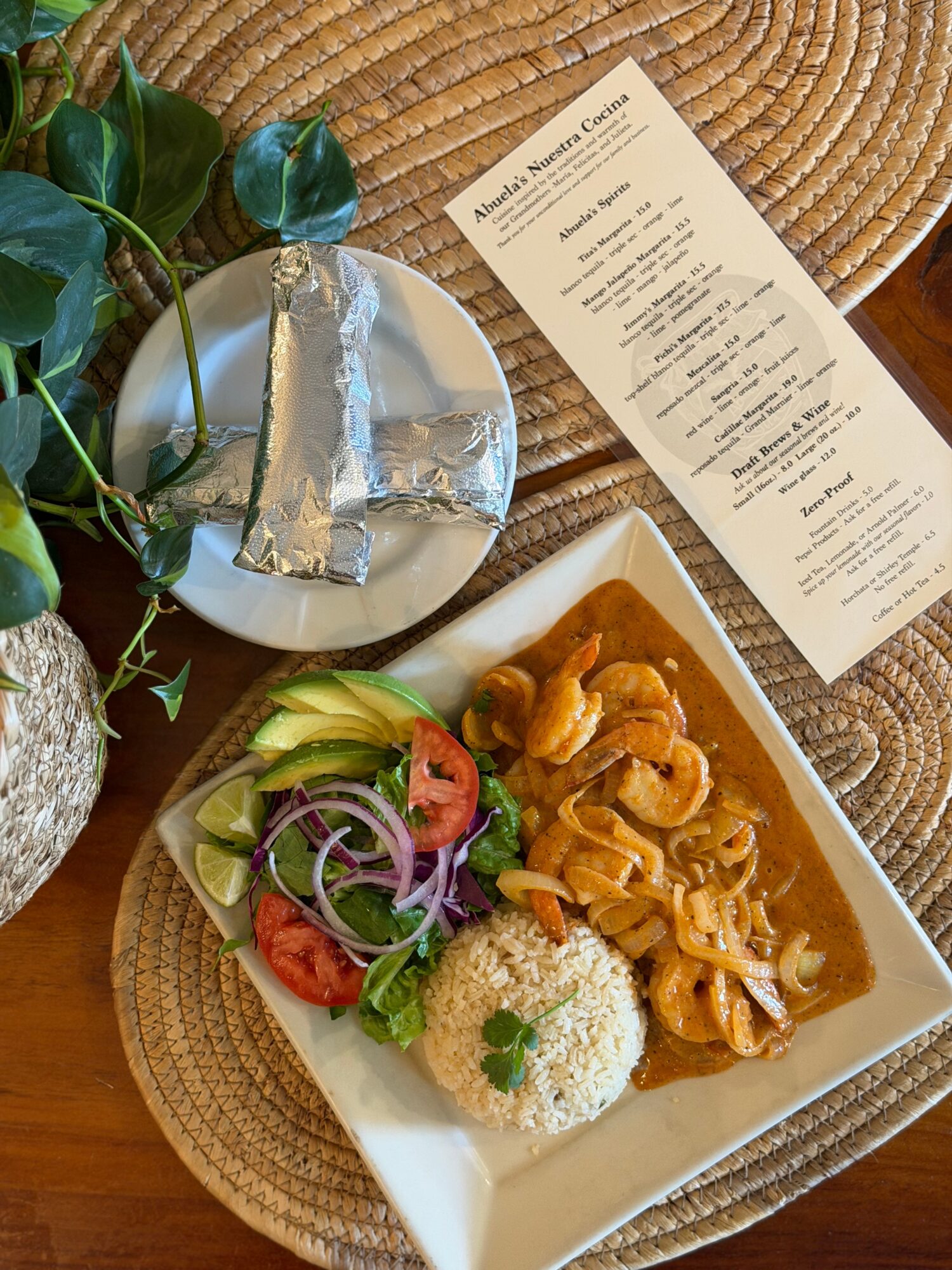 Plate with rice, vegetables, and curry, a menu, foil-wrapped items, and a plant on woven placemats.