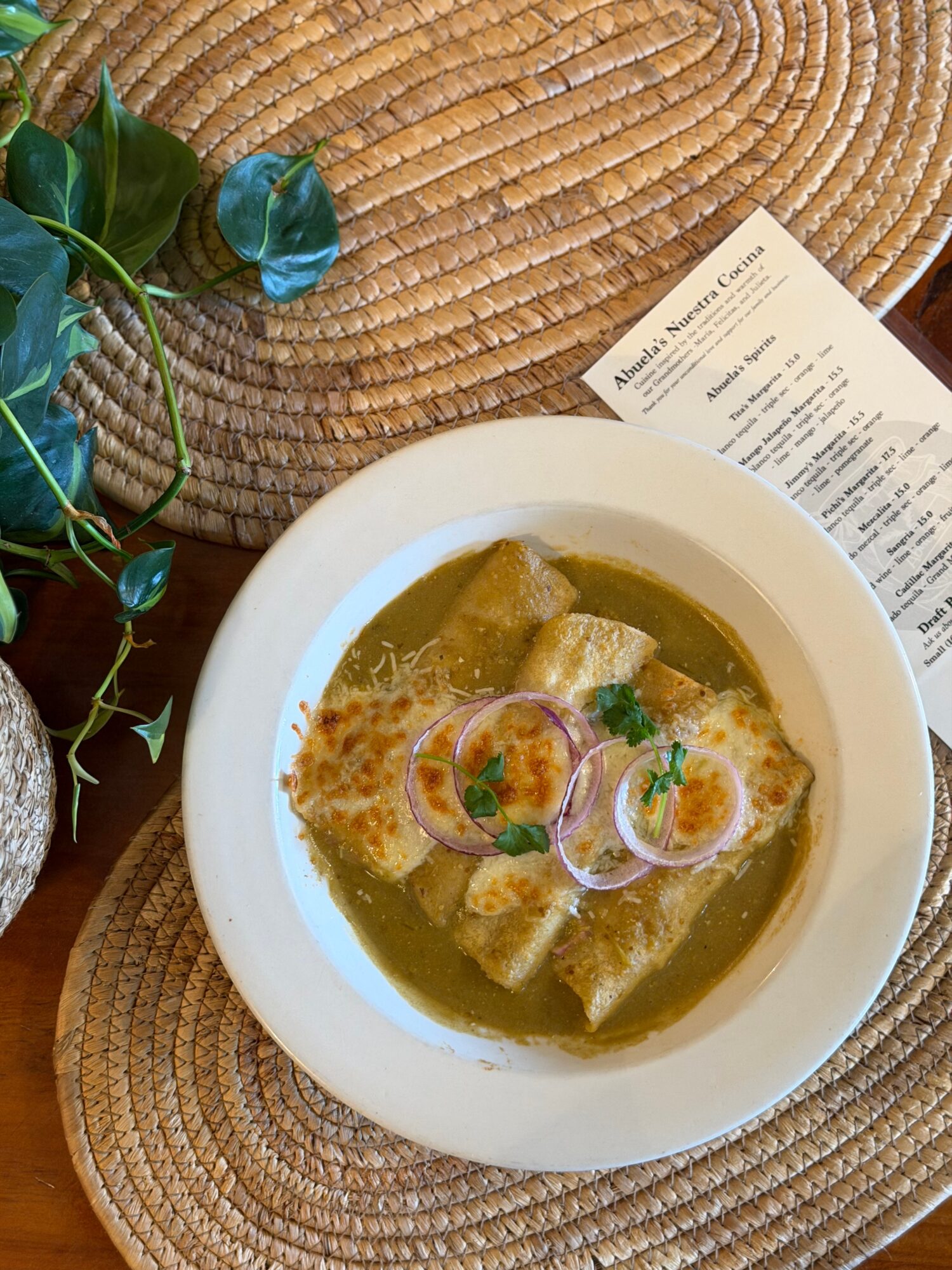 Bowl of soup with herbs and bread on a woven placemat, with a menu and plant nearby.