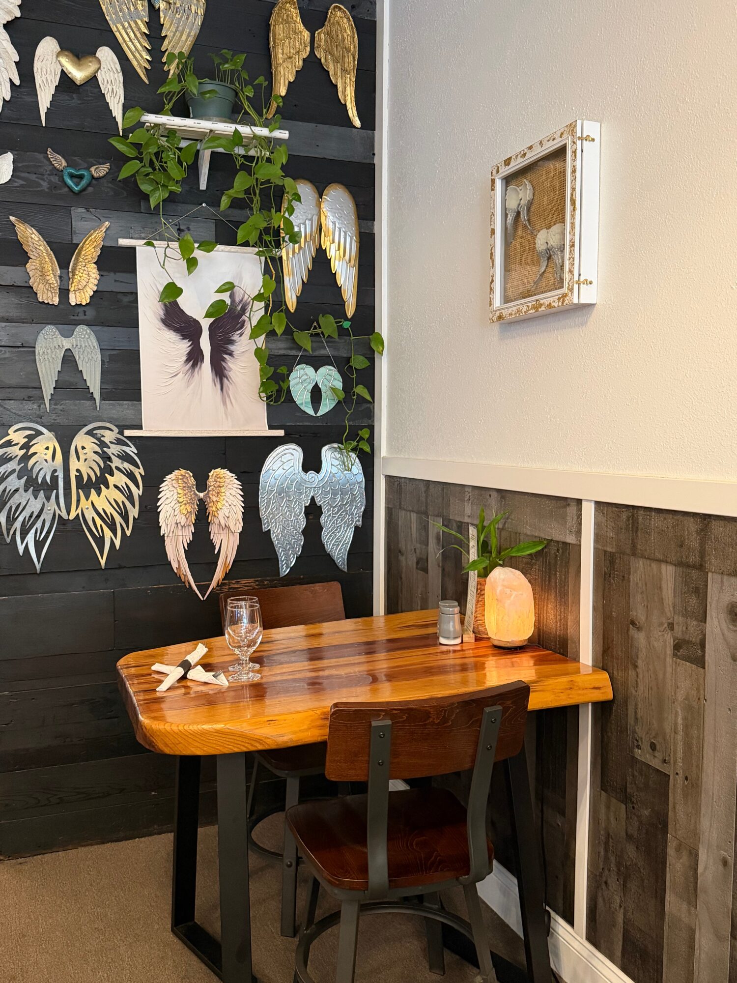 Wooden table with a glass, a candle, and a small plant, against a wall with butterfly artwork and a framed mirror.