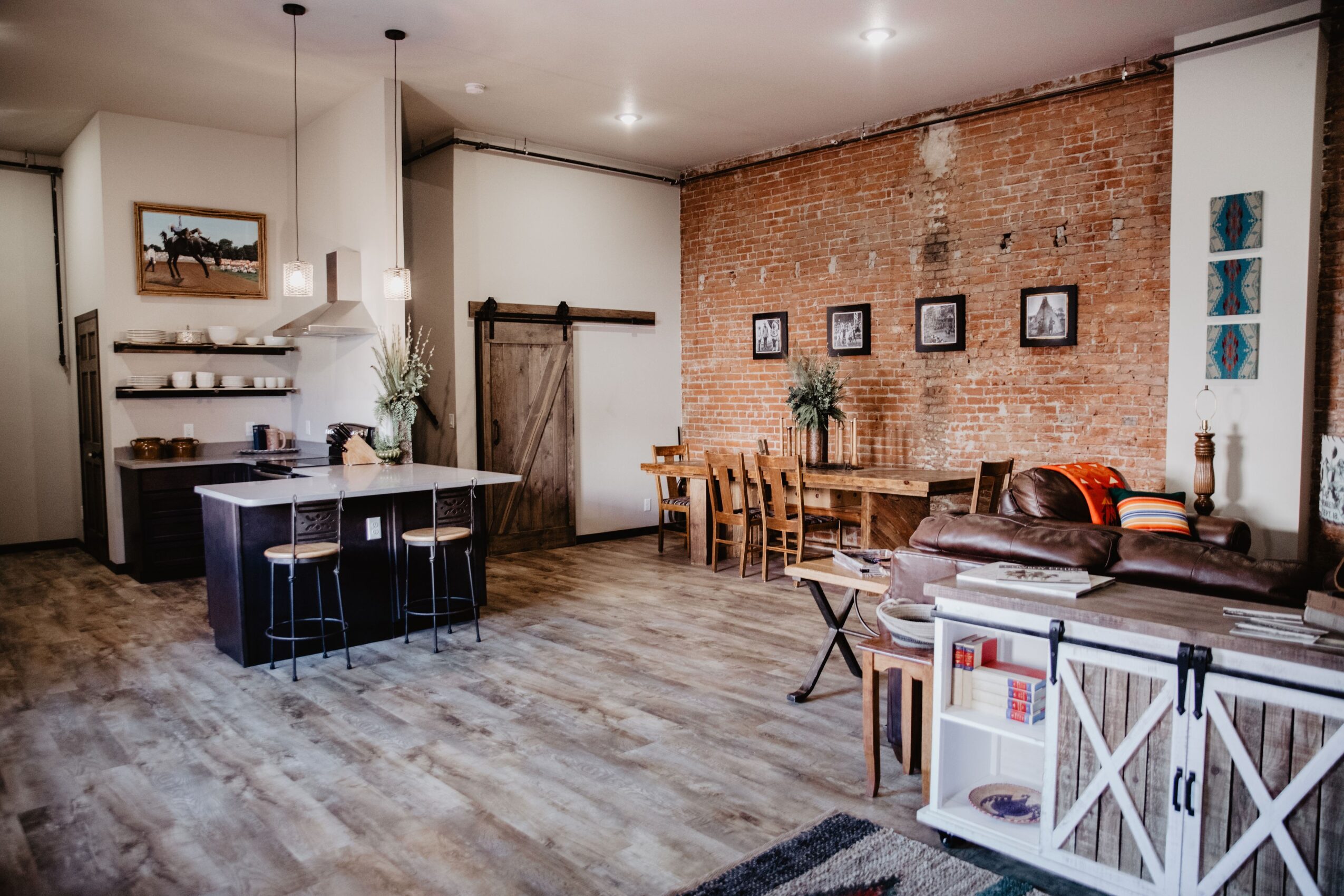 Living room with brick wall, wooden furniture, and a kitchen island with stools, decorated with pictures and plants.