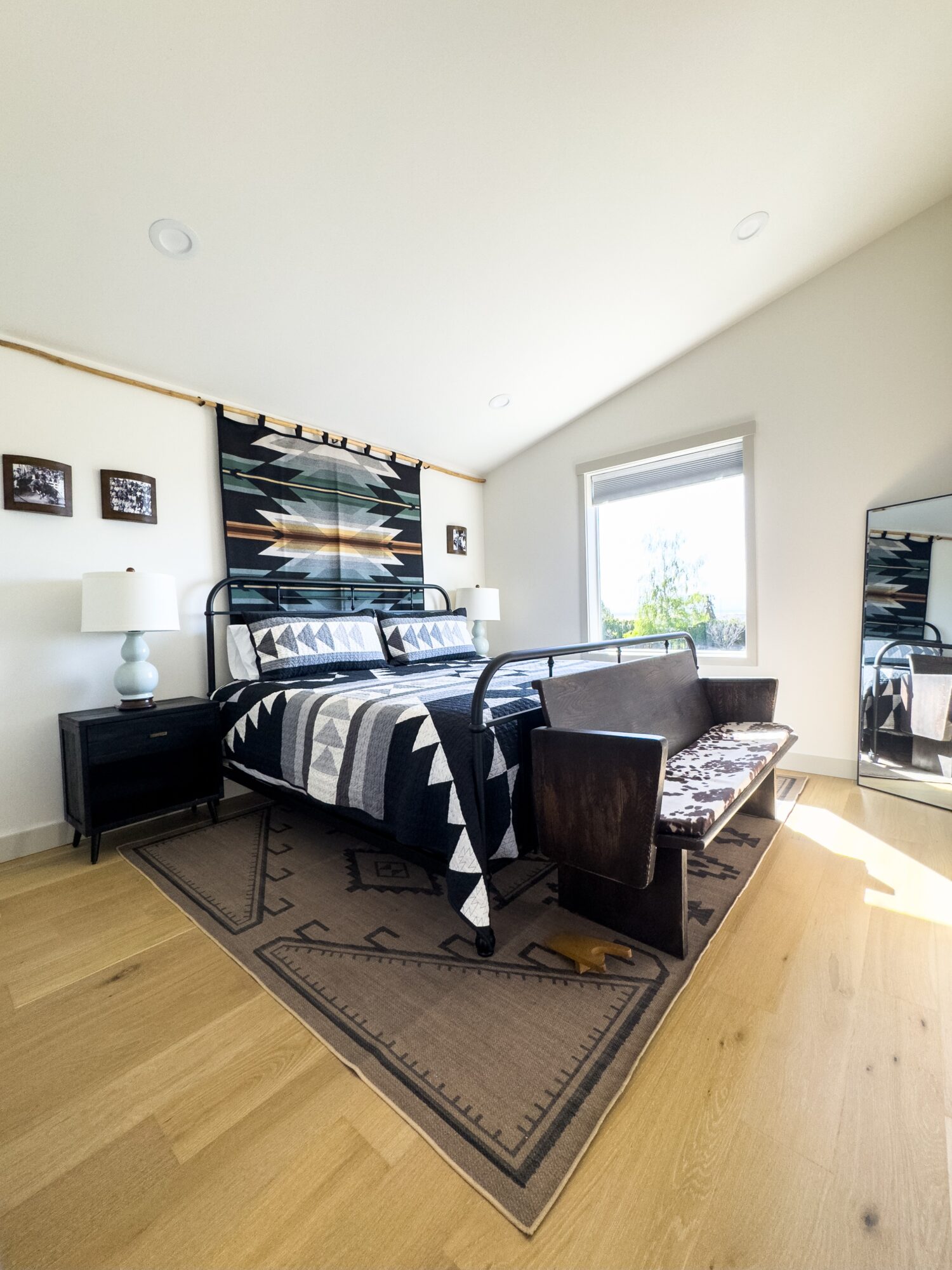 Bedroom with bed, nightstands, lamps, window, and mirror, sunlight streaming in, wooden floor, and patterned wall hanging.