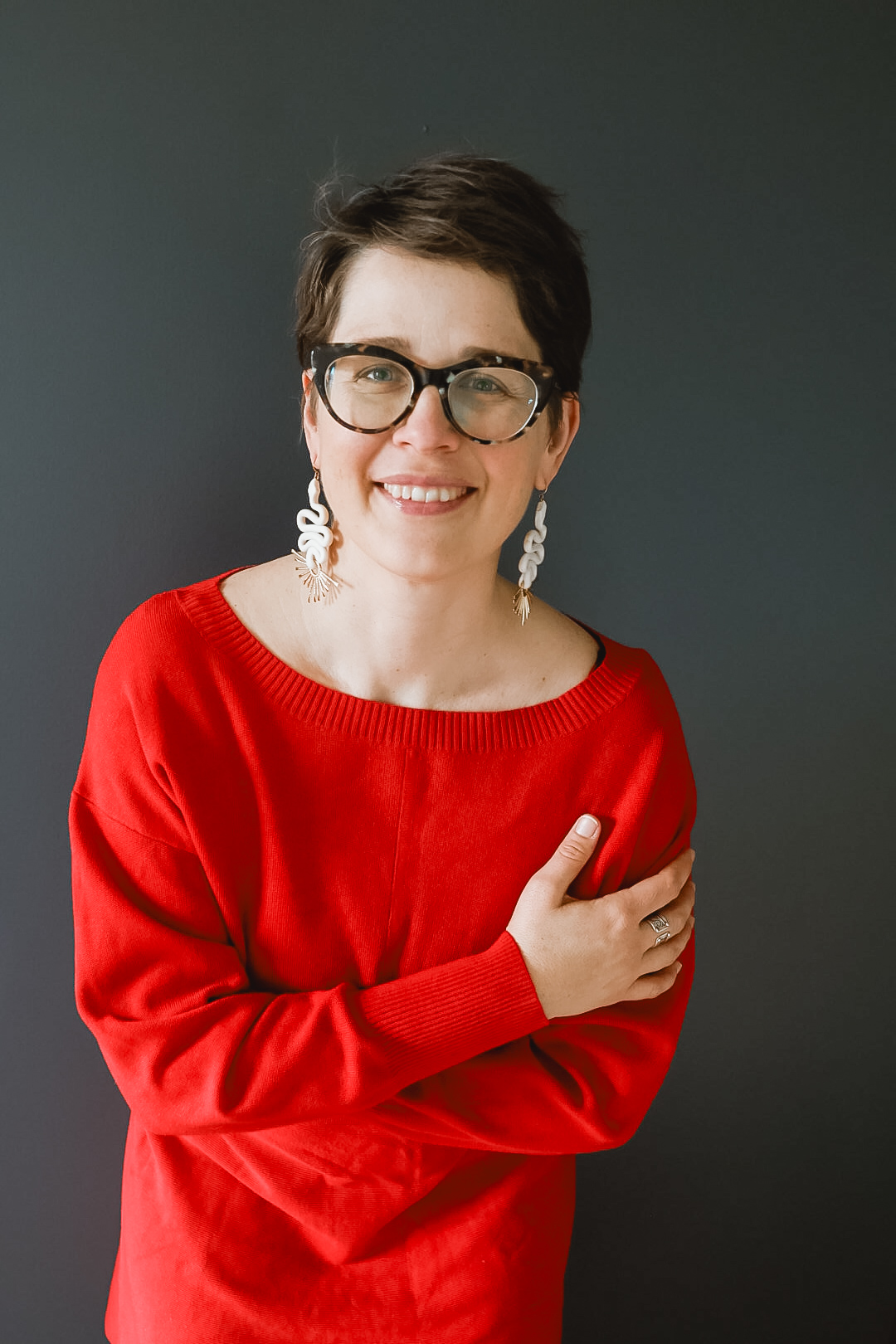 Woman with short dark hair, glasses, and earrings, smiling, wearing a red top, against a dark background.