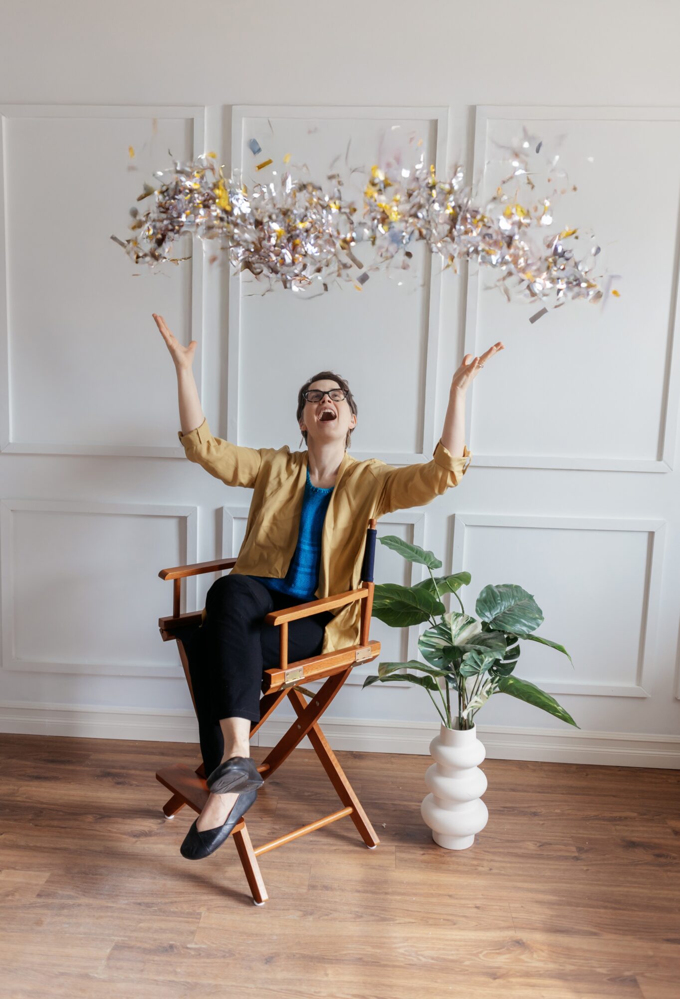 Person sitting on a wooden chair with arms raised, throwing confetti in a room with white walls and a potted plant.