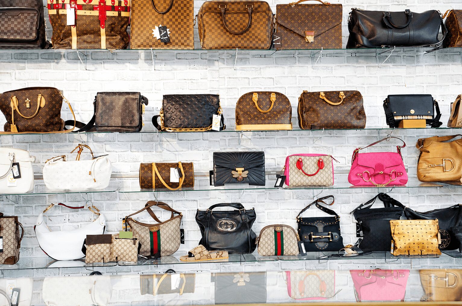 Display of various handbags on shelves against a white brick wall, arranged in rows with different styles and colors.