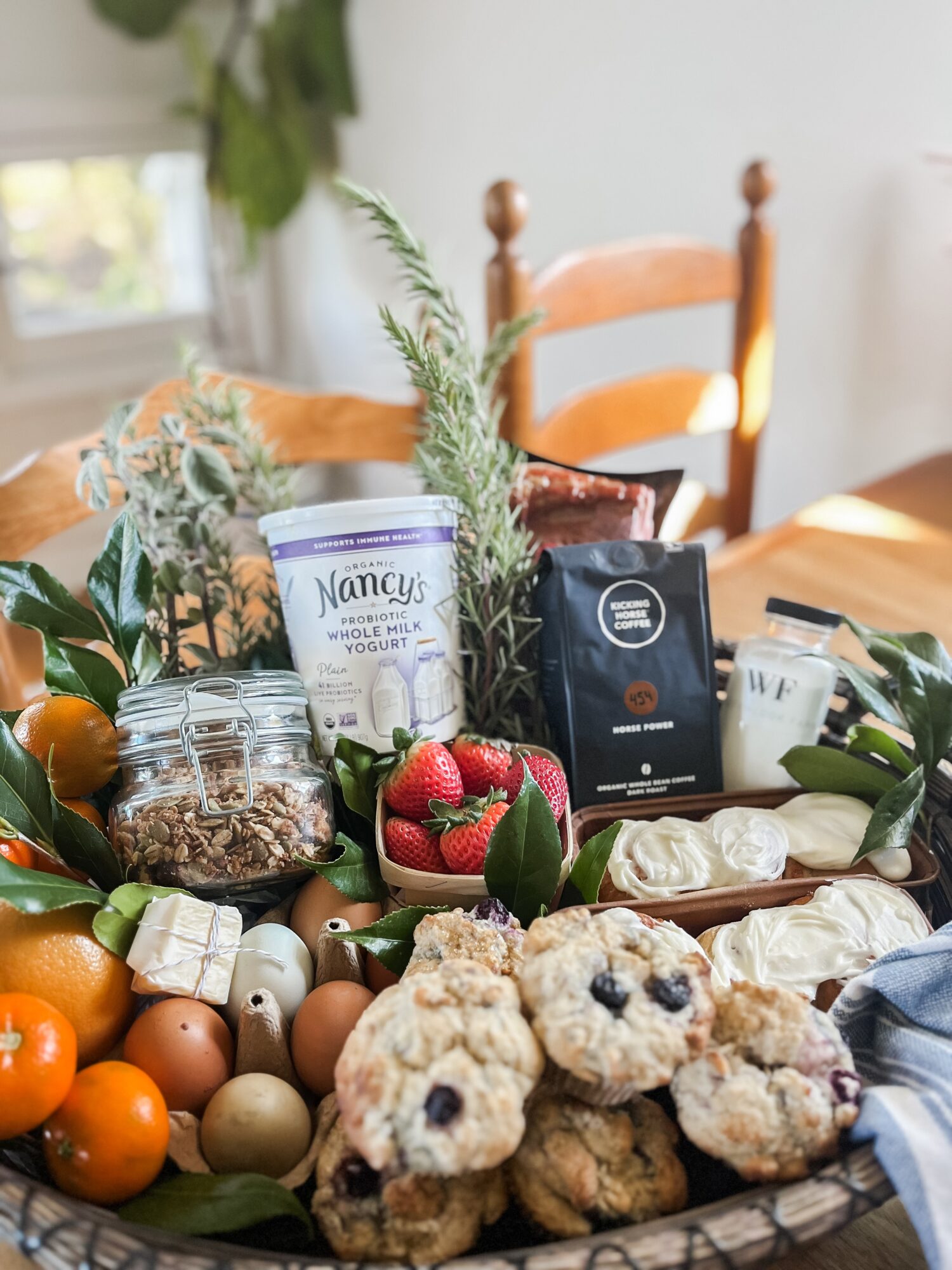 Assorted baked goods, strawberries, oranges, and packaged food items on a table with chairs in the background.