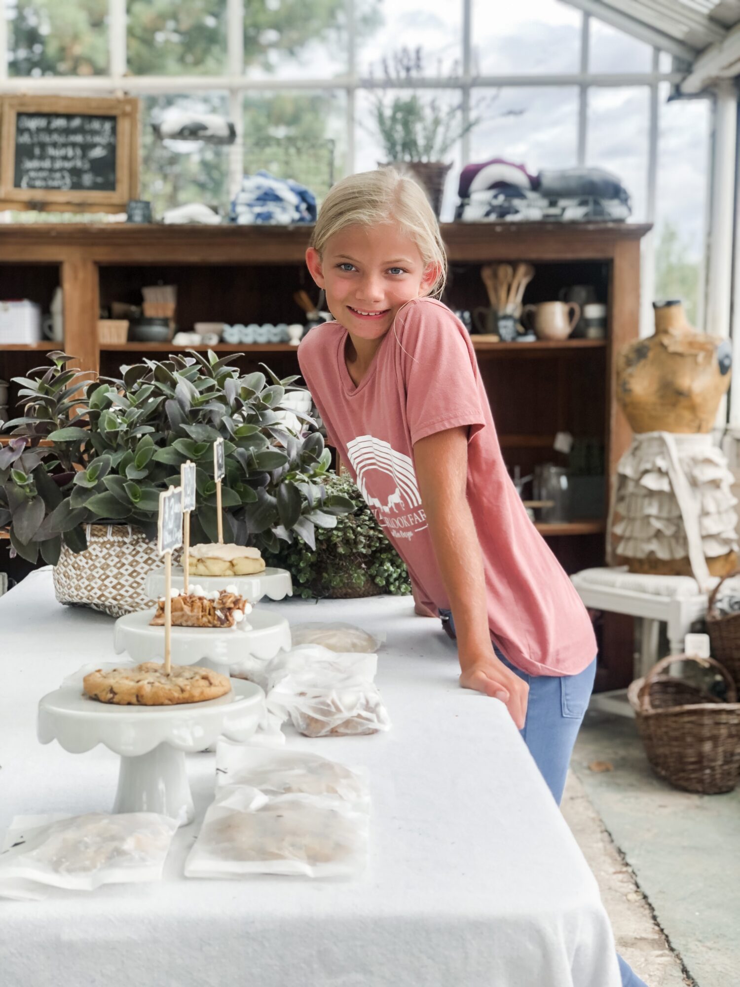 Smiling girl leaning on a table with baked goods in a bright, indoor setting with plants and shelves.