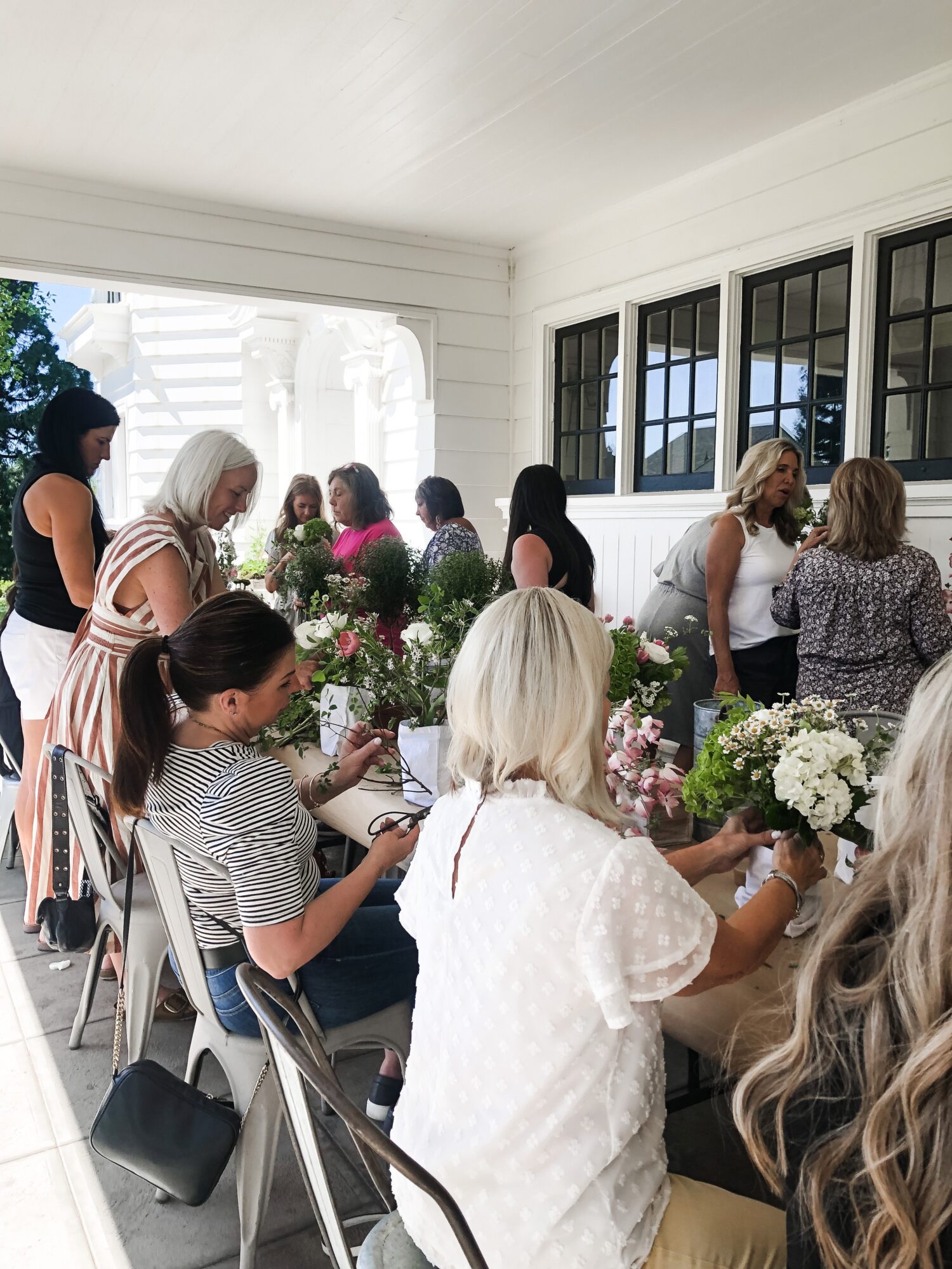 Group of women gathered indoors, some seated and some standing, with flowers and windows visible, engaged in conversation.