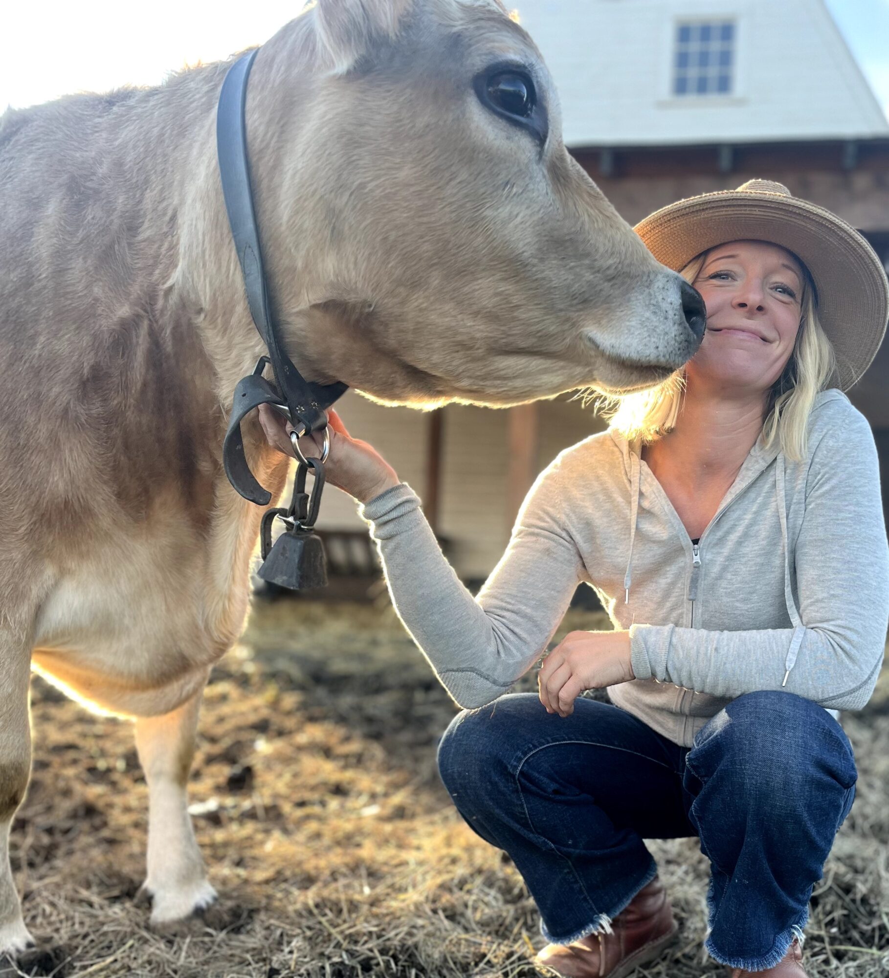 Woman in hat crouching next to a horse, touching its nose, outdoors with a barn in background.