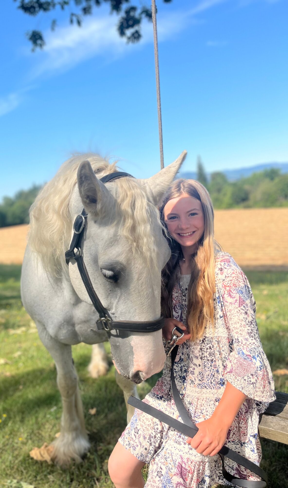 Young woman with long hair smiling next to a white horse outdoors on a sunny day.