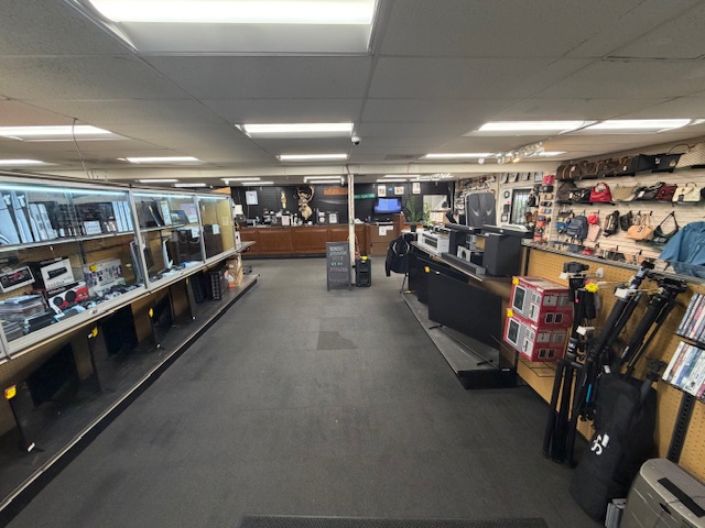 Empty store aisle with shelves of electronics and accessories, checkout counters at the back, and a gray carpeted floor.