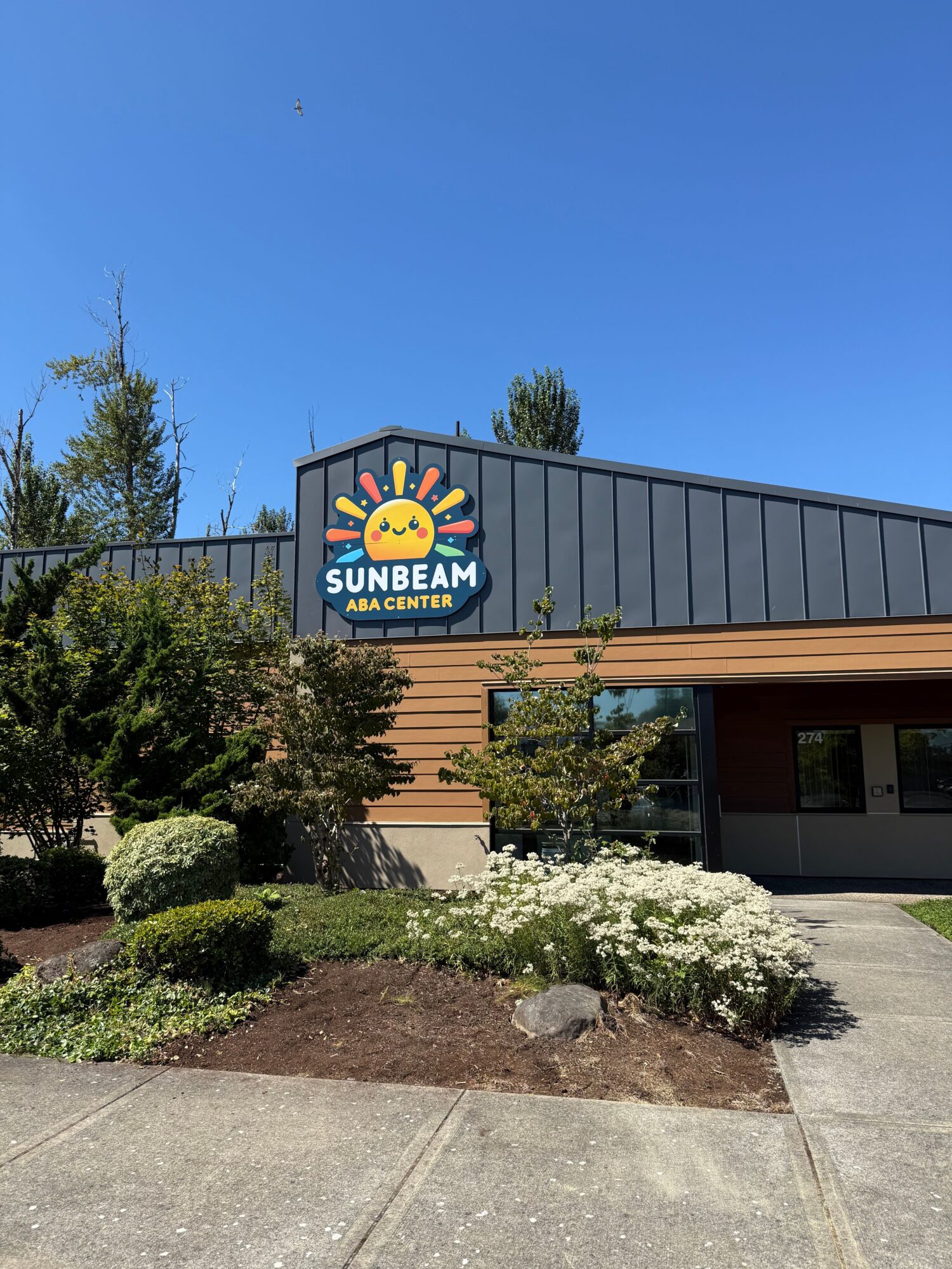 Sunshine Sub Shop sign outside a building with plants and a sidewalk, under a clear blue sky.
