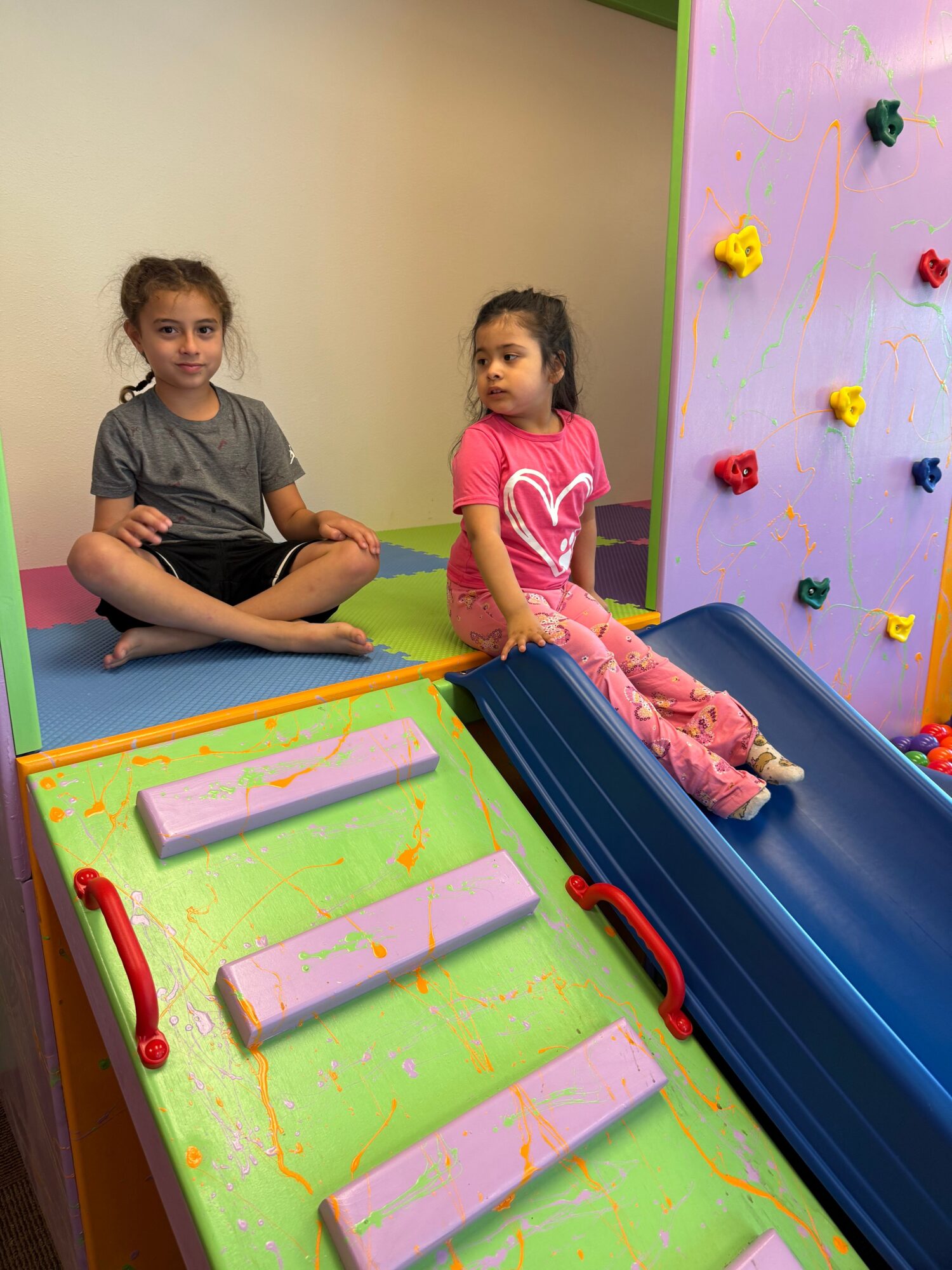 Two children playing on a colorful indoor slide, one sliding down and the other sitting at the top, with a pink and green background.