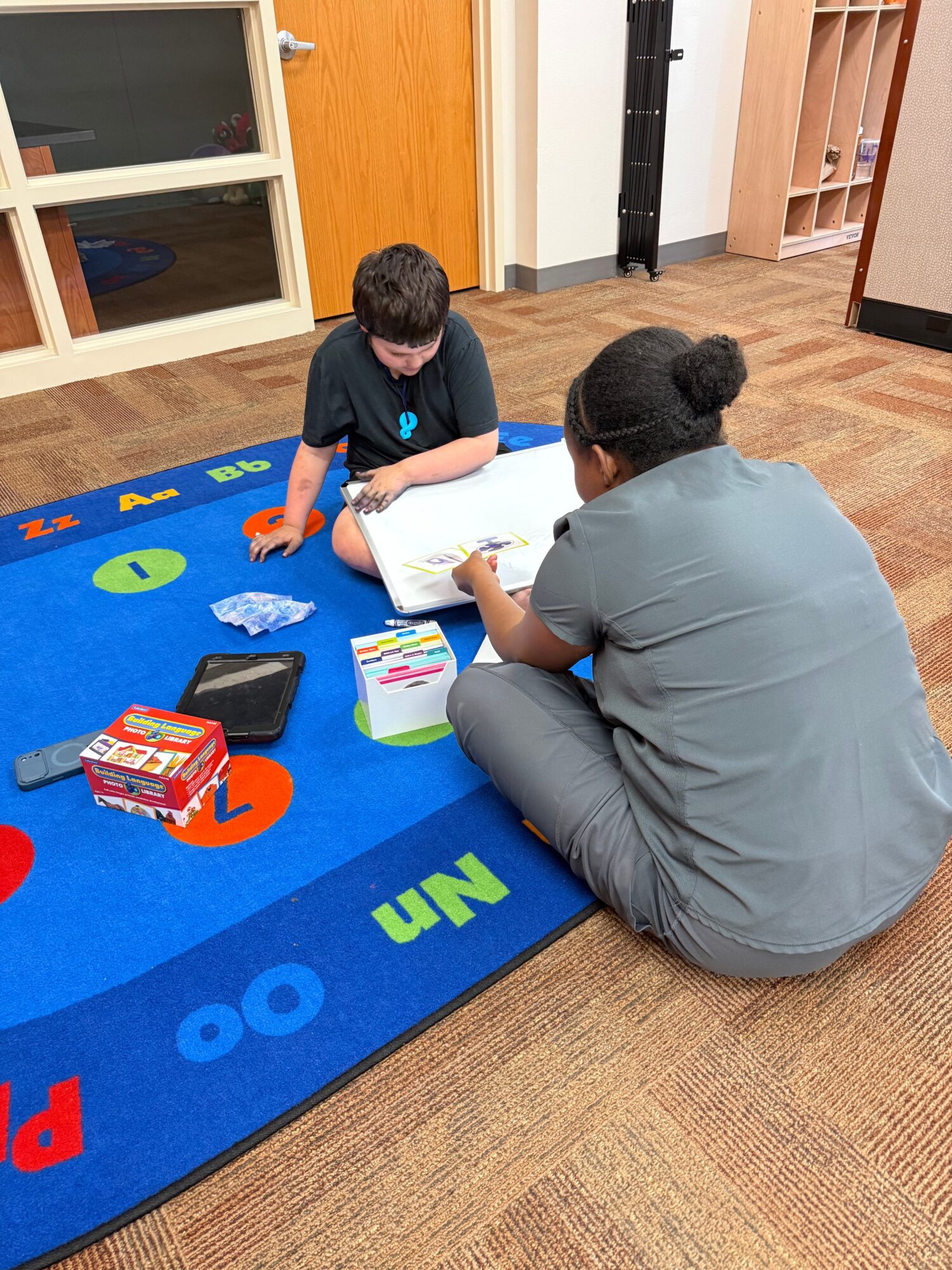 Two people sitting on a blue educational mat, engaging with a tablet and educational materials, in a room with wooden floors and furniture.