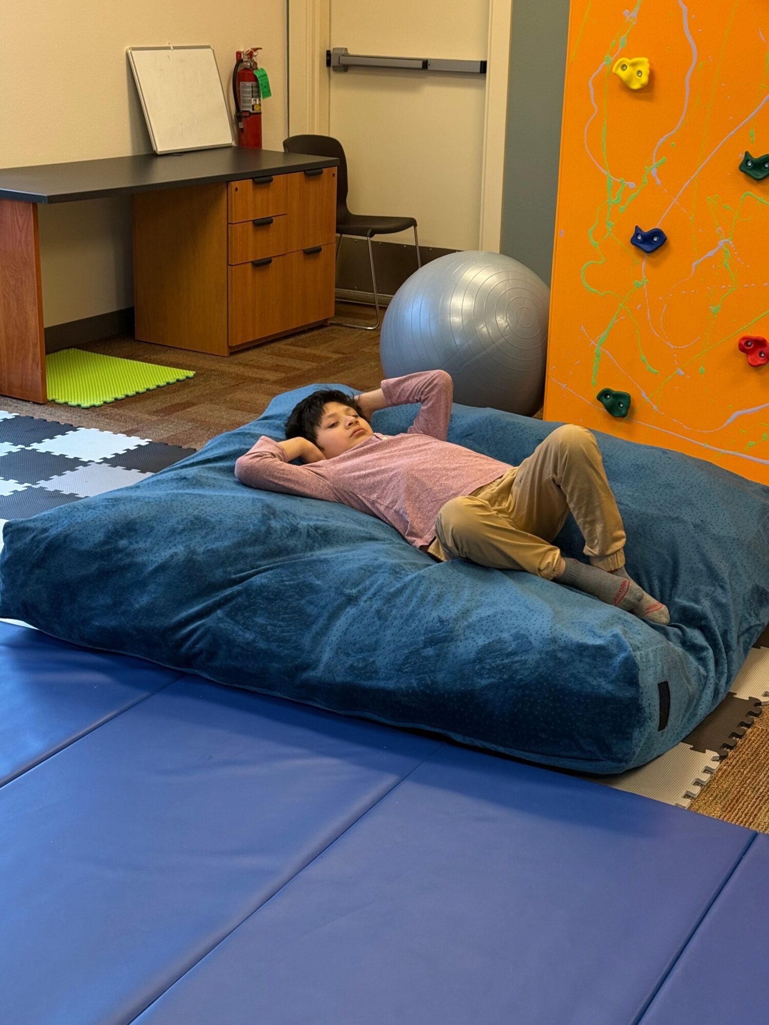 Person lying on a blue bean bag in a room with colorful climbing wall, exercise ball, and foam mats.