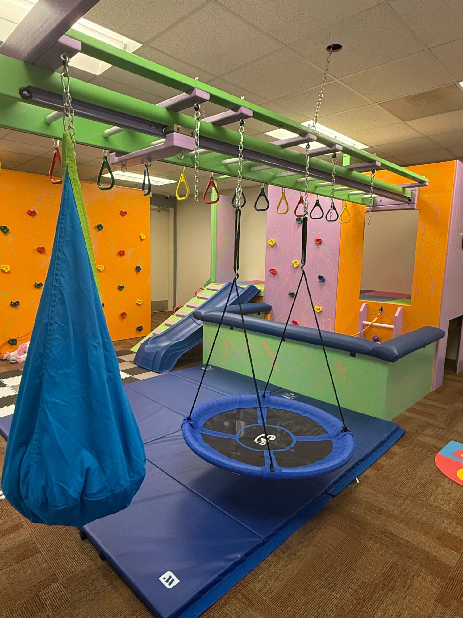 Indoor play area with colorful equipment, a blue mat, and a blue fabric cone, on a wooden floor.