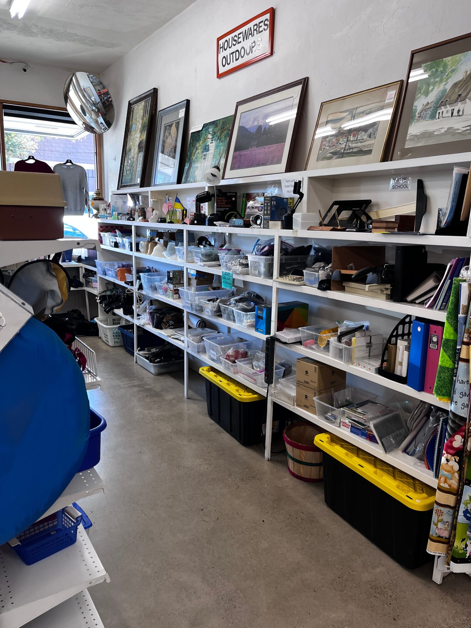 Shelves filled with various items, framed pictures on the wall, and a person in a blue shirt in a room.