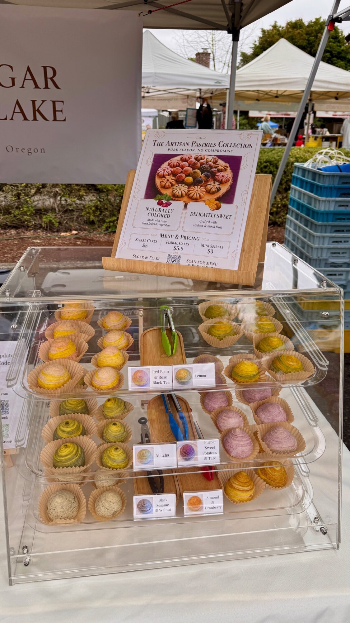 Display of colorful macarons in a clear case at an outdoor market, with a menu and tents in the background.