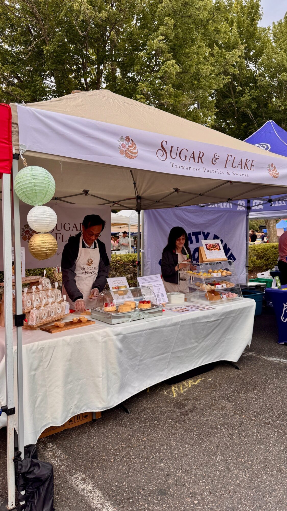 People behind a table at a booth with a white canopy, selling baked goods outdoors, trees in background.