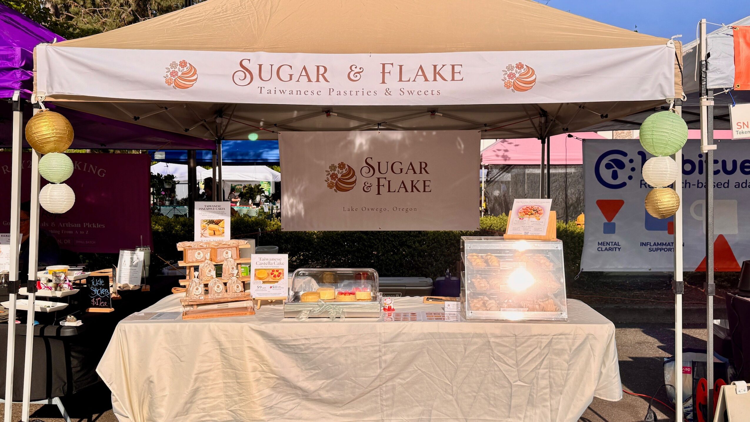 Market stall with a beige canopy and a table covered with a beige cloth, displaying baked goods and decorations.