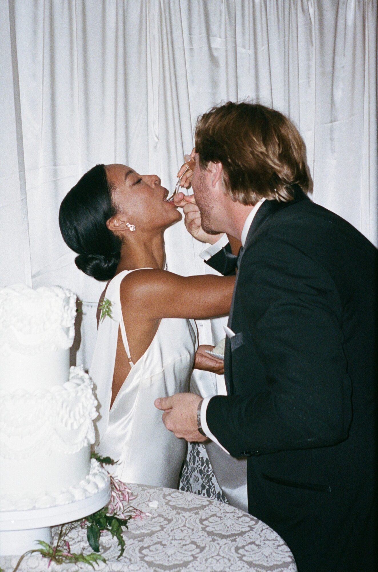 Two people in formal attire sharing a cake slice at a celebration, with a white curtain backdrop and a decorated table.