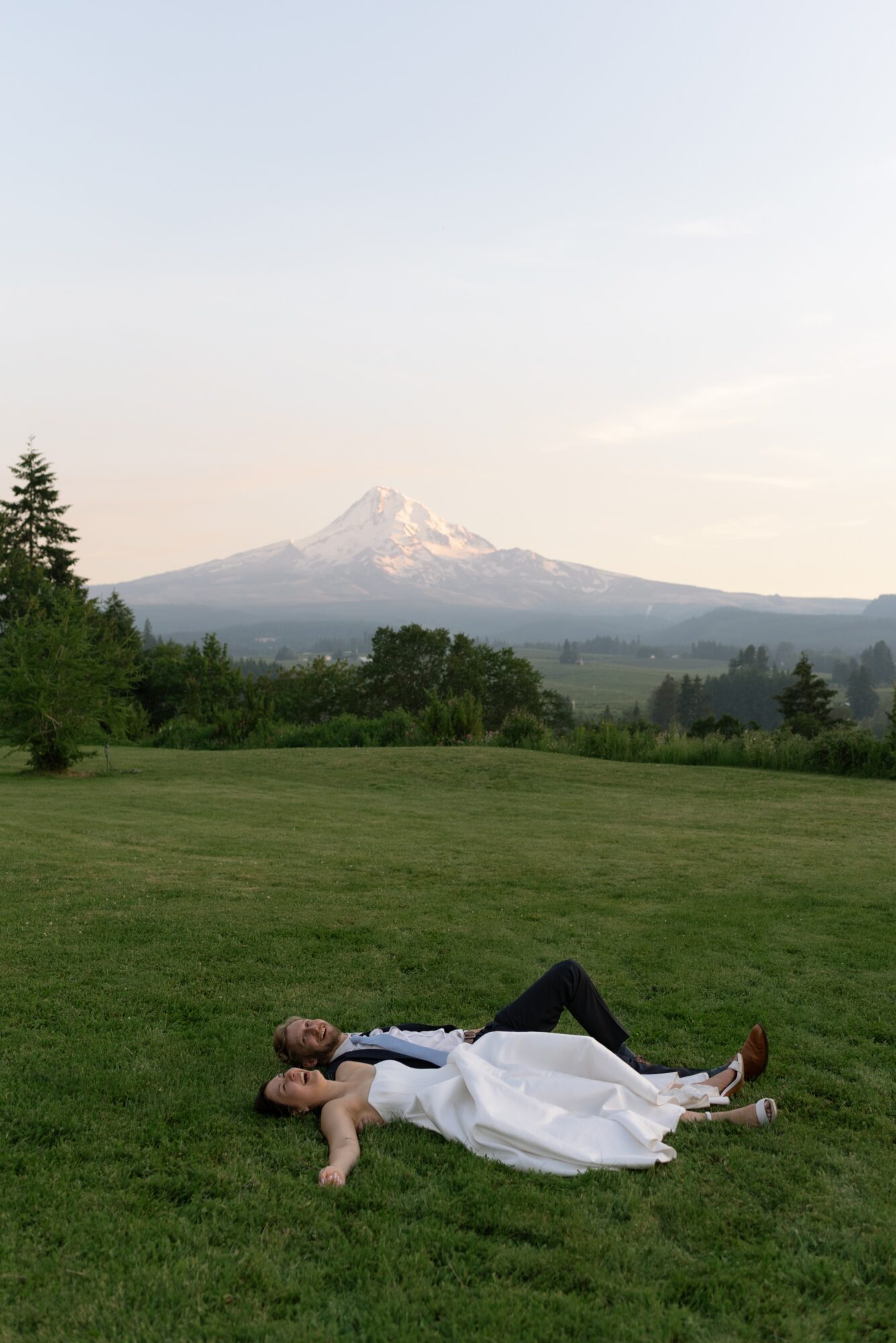 Person lying on grass in a white dress with a mountain in the background, trees, and a clear sky.