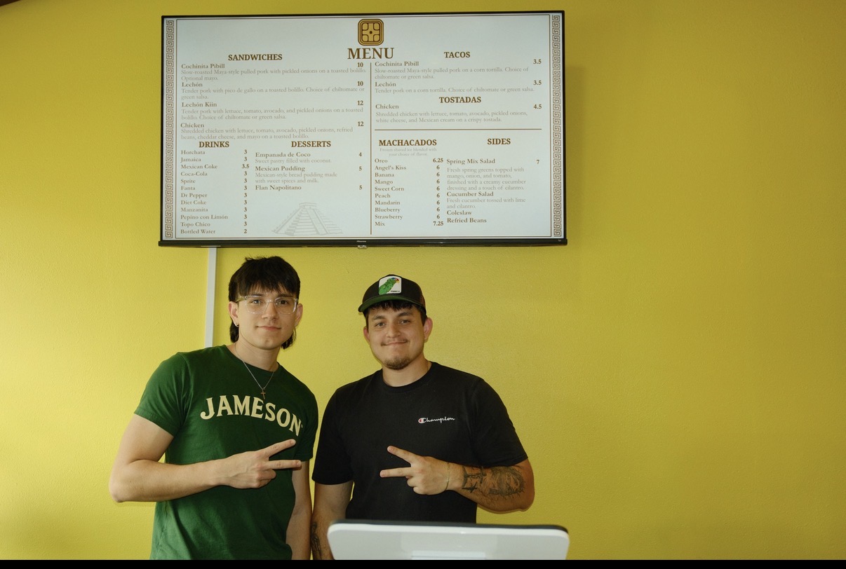 Two young men stand in front of a yellow wall with a menu screen above them, both making peace signs with their hands.