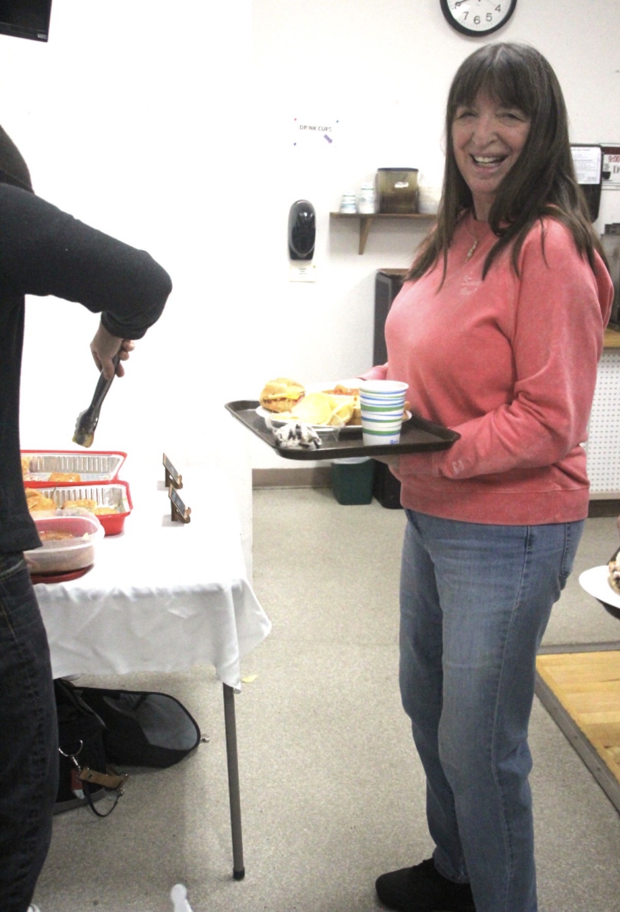 Woman in pink sweater holding a tray with food, smiling, in a room with a clock and shelves, standing near a table.
