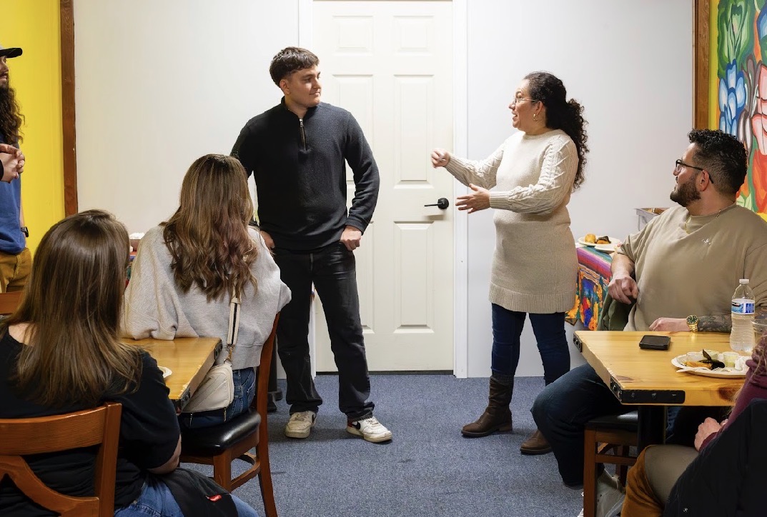 Group of people in a room, woman speaking to a young man, others seated at tables, colorful artwork on wall.