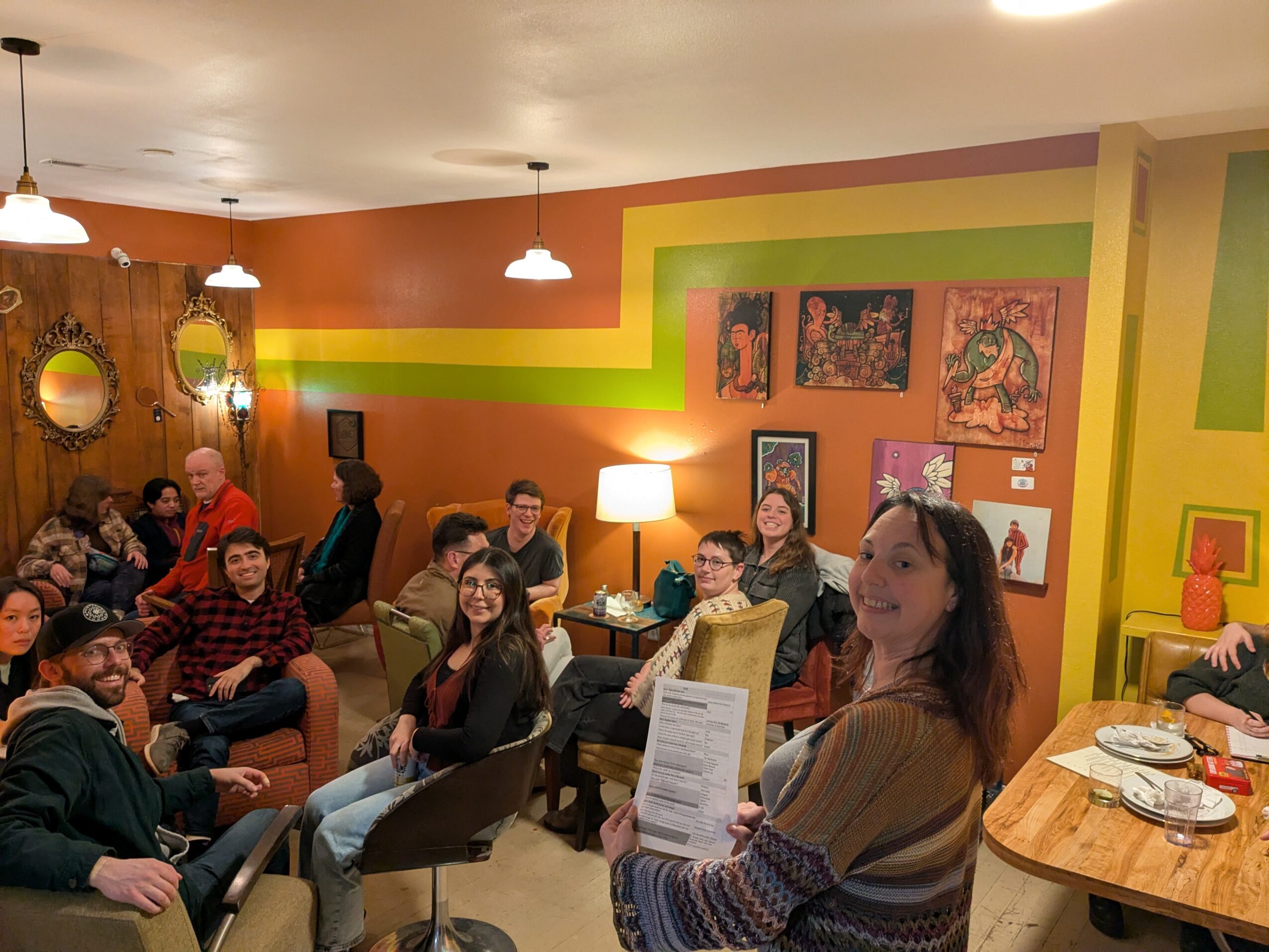 Group of people sitting and standing in a colorful room with paintings, a lamp, and a table with plates.