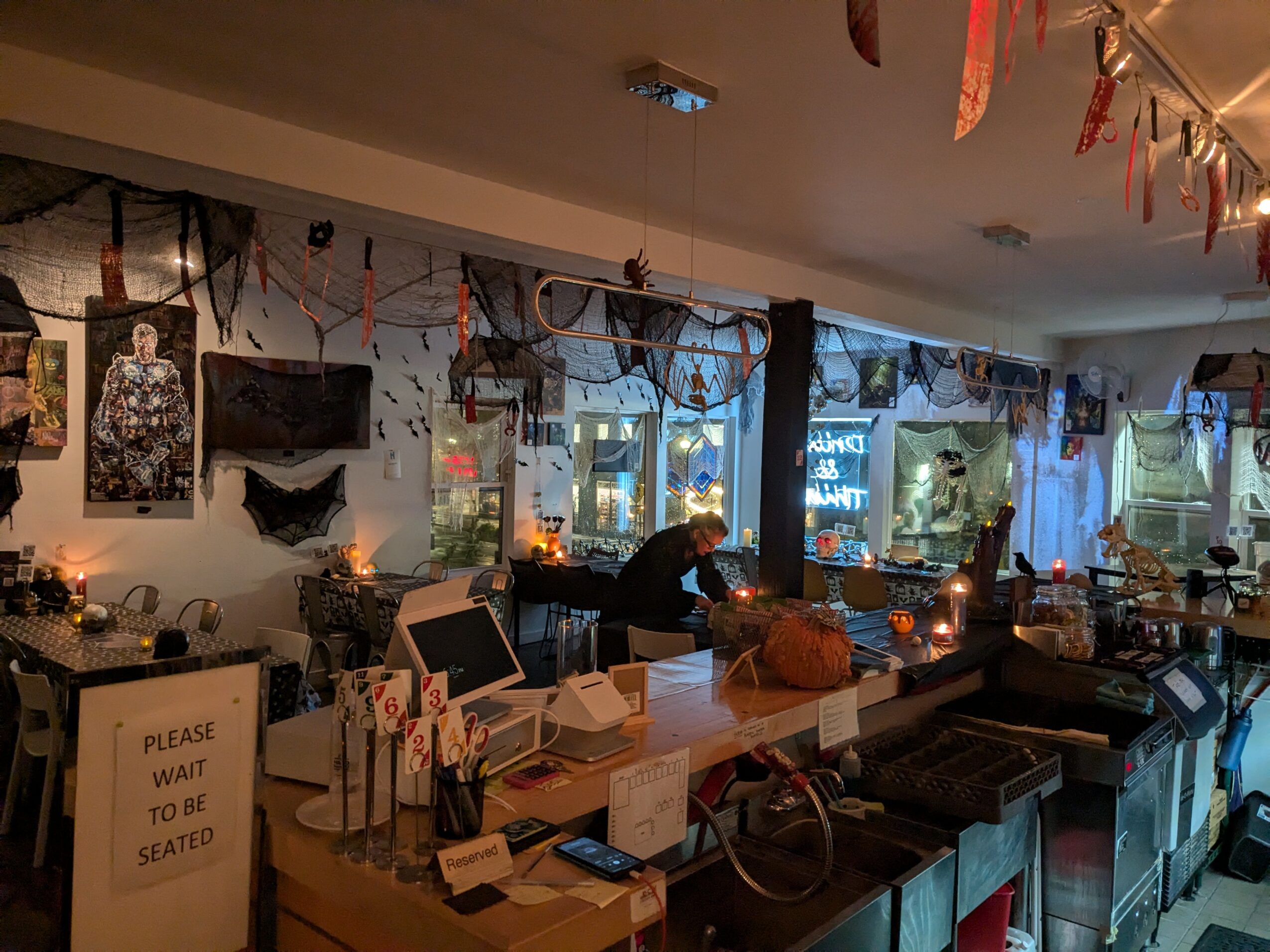 Indoor space decorated with hanging banners, lights, and Halloween-themed decorations, with a person sitting at a counter near windows.