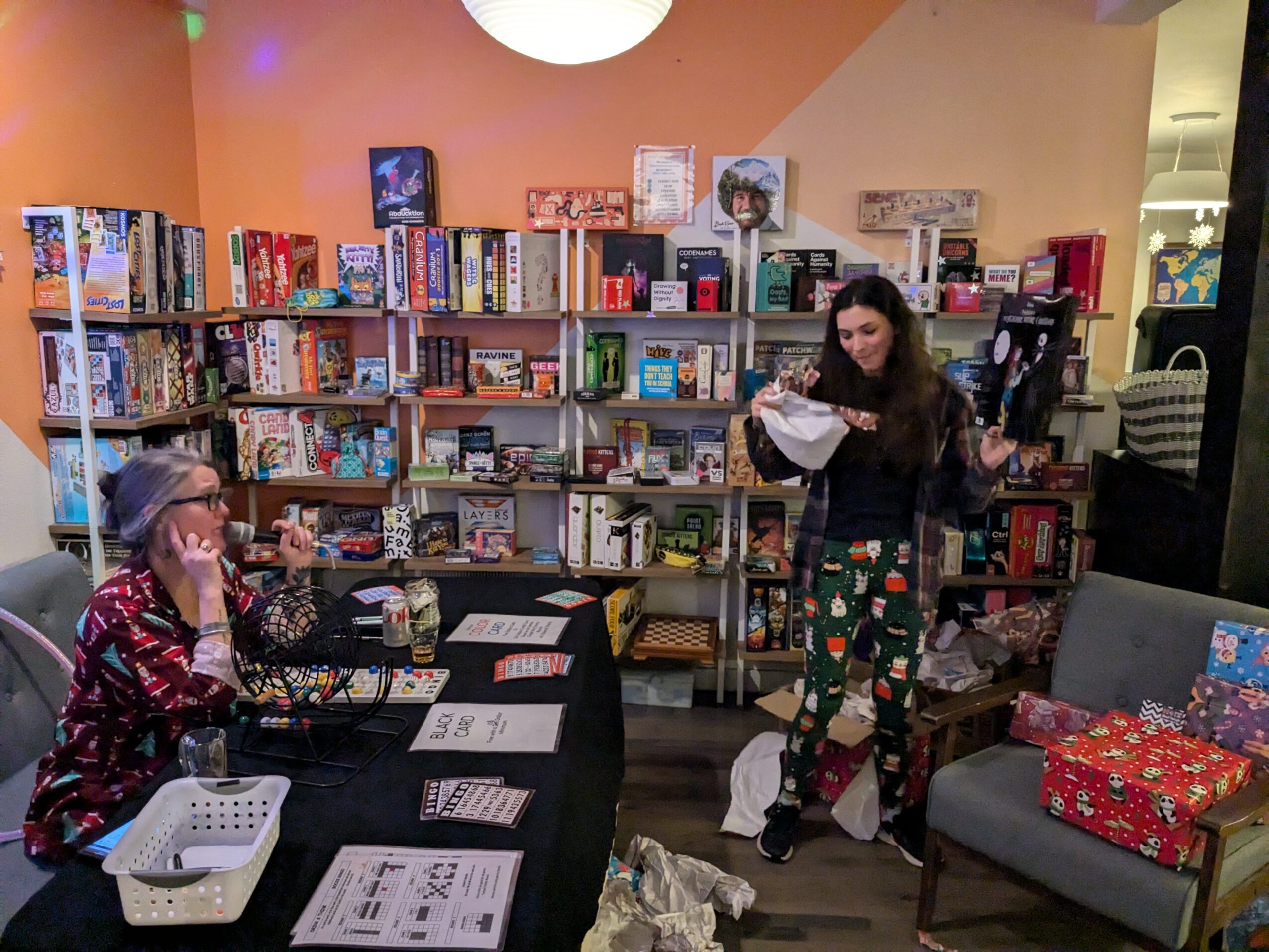 Two women in a bookstore, one sitting at a table with books and the other standing reading a paper, bookshelves in background.