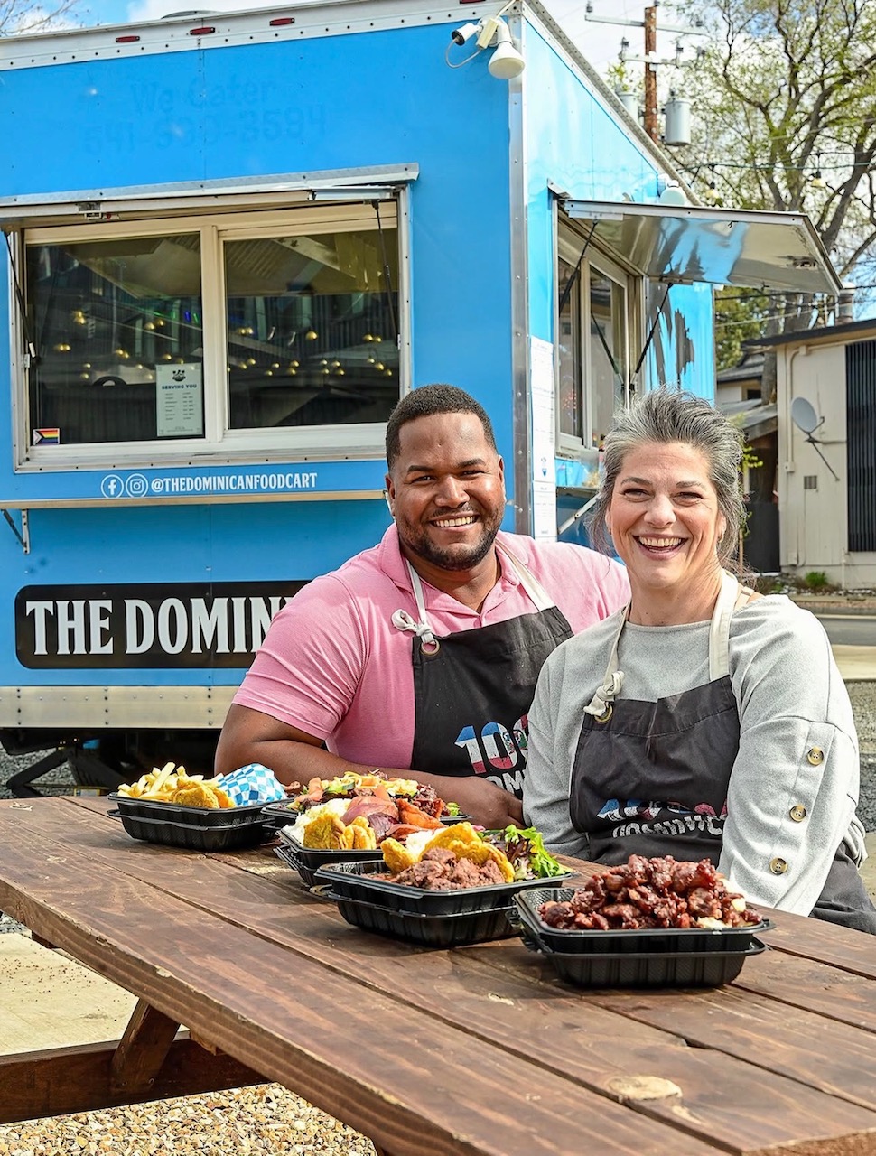 Two people smiling behind a food truck with dishes on a wooden table outdoors.