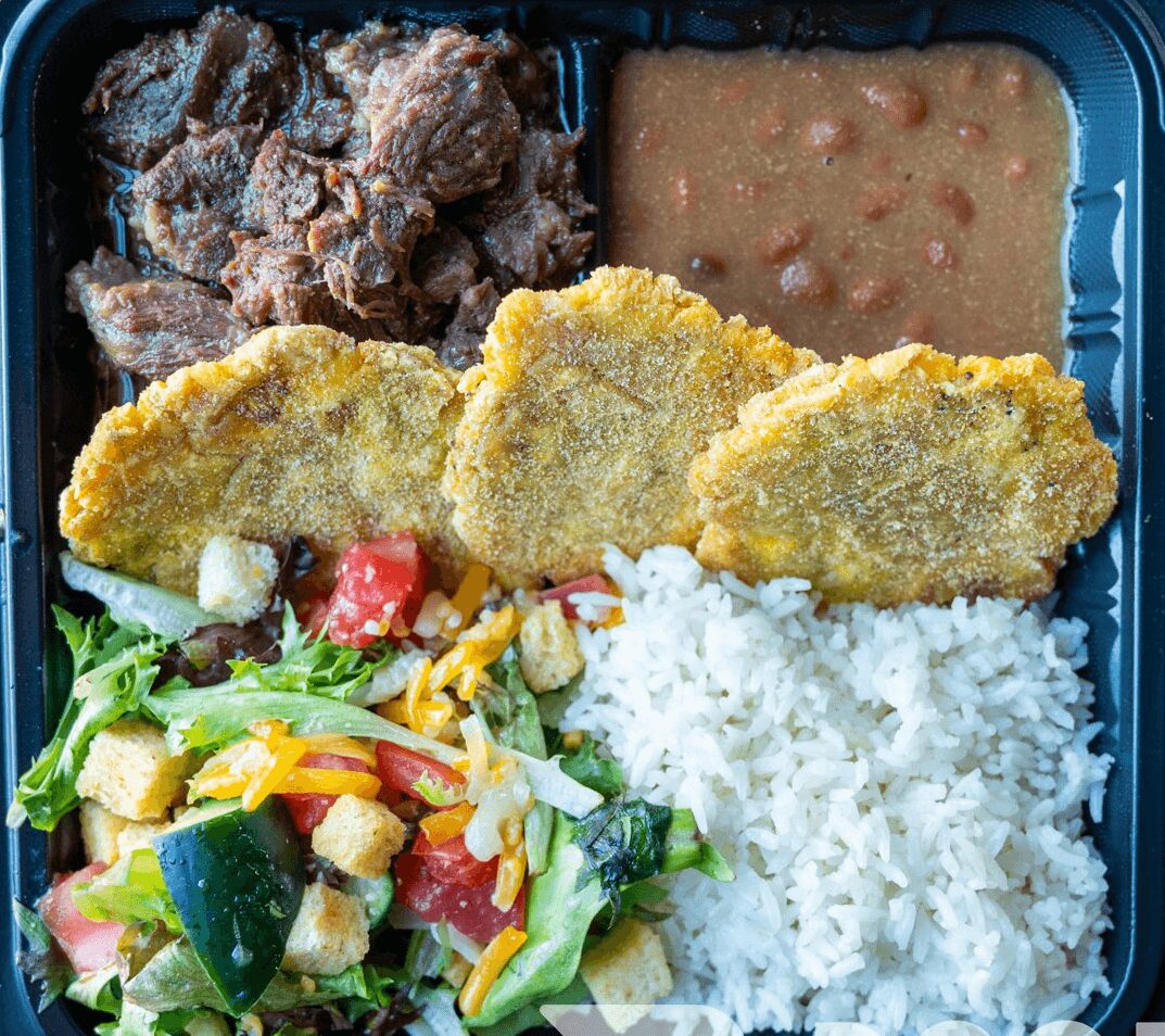 Fried chicken pieces, rice, vegetable salad, beans, and fried plantains in a black tray.