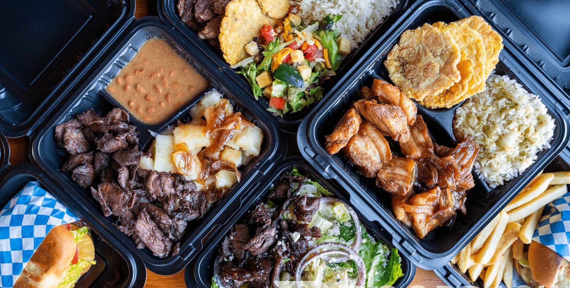 Four black meal containers with fried chicken, salad, rice, and fries, on a table with a checkered napkin.