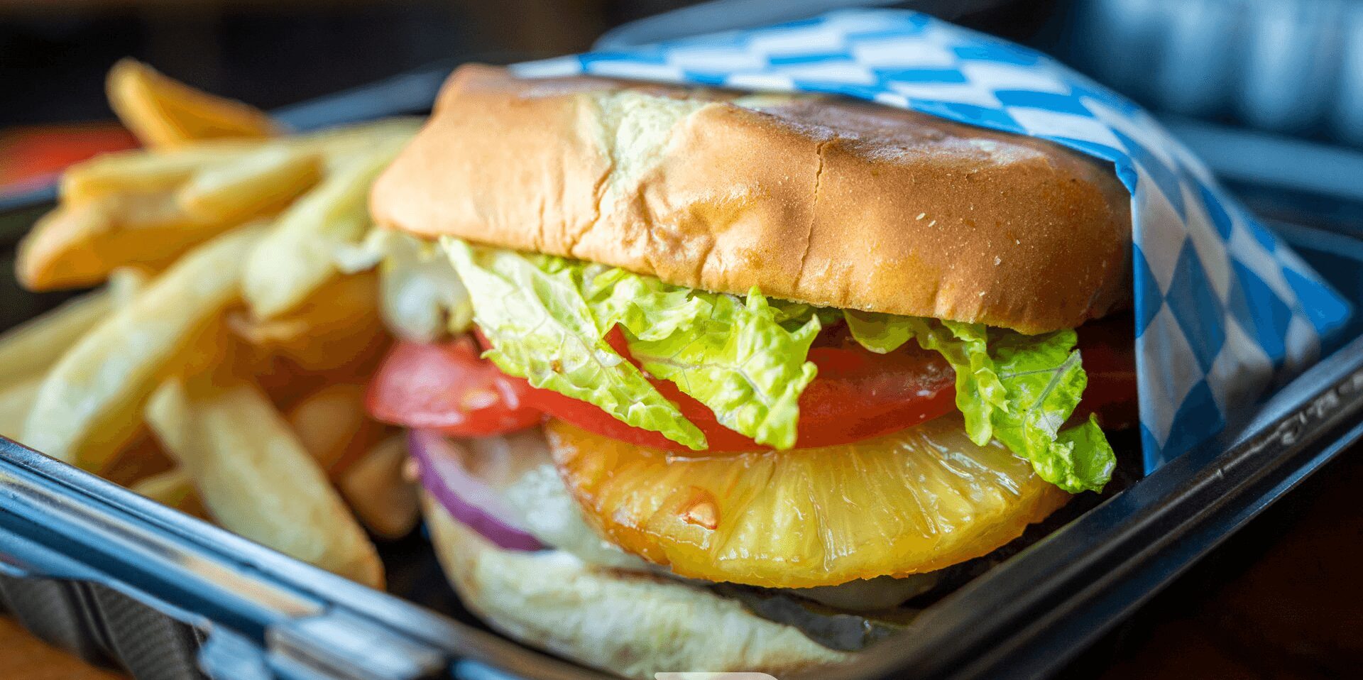 A burger with lettuce, tomato, onion, pineapple, and fried chicken in a bun, served with fries on a tray.