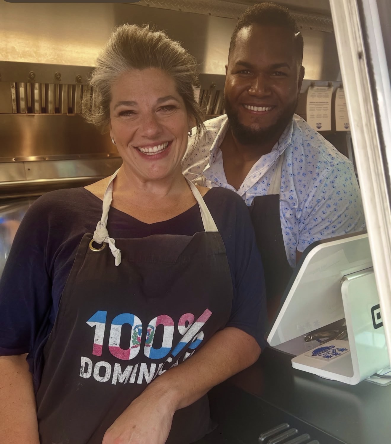 Two smiling people, one older woman and one younger man, in a kitchen setting, wearing aprons, standing close together.
