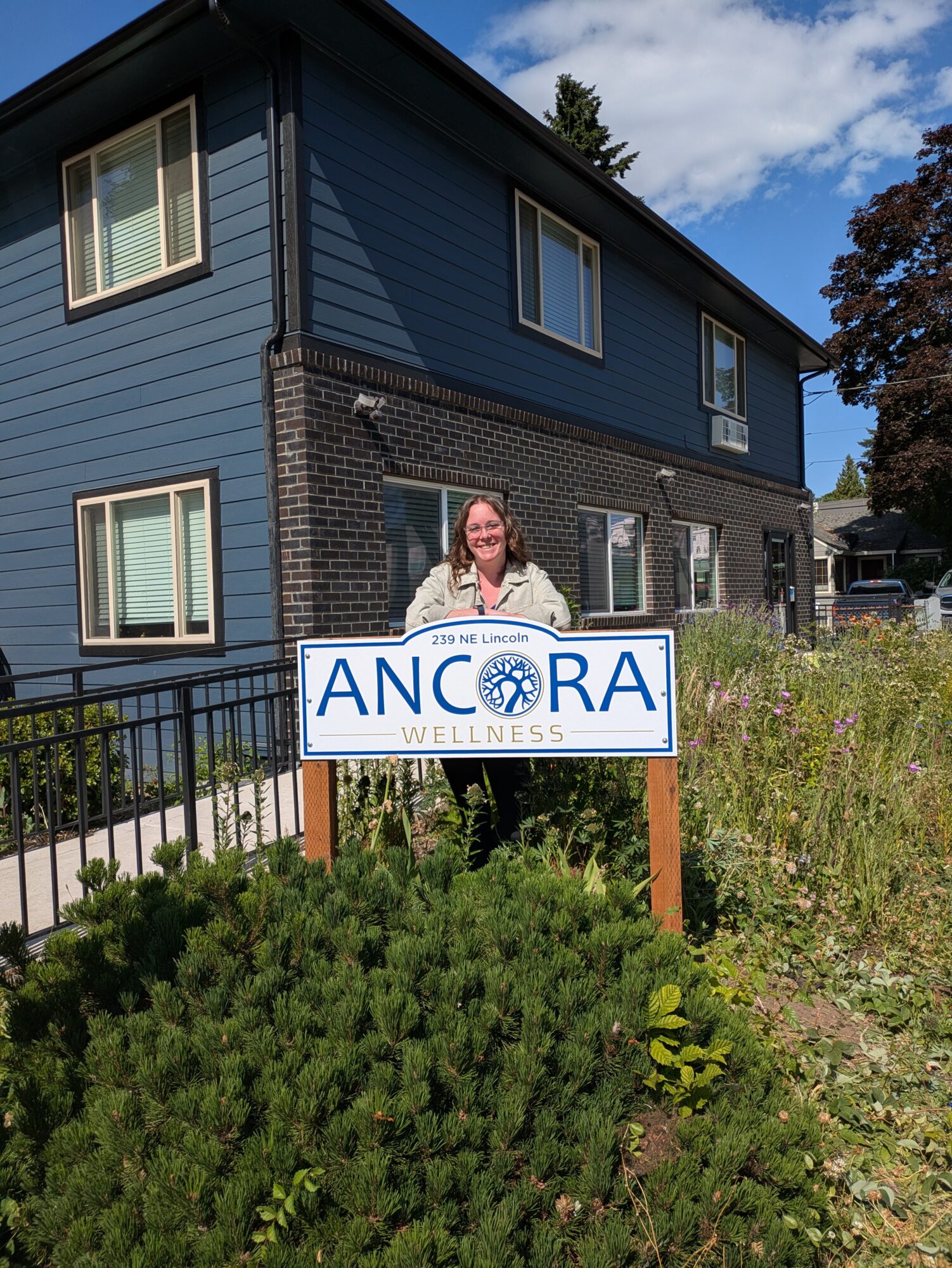 Woman standing outdoors behind a sign for Ancora Wellness, in front of a blue house with windows, greenery, and a partly cloudy sky.