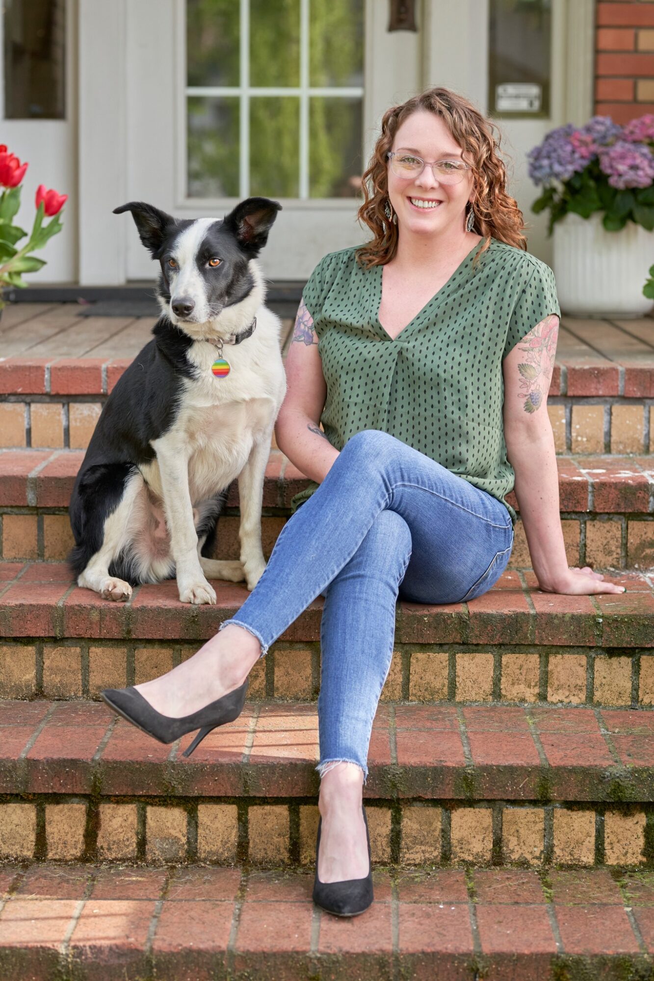 Woman with curly hair and glasses sitting on brick steps with a black and white dog, in front of a house with flowers.