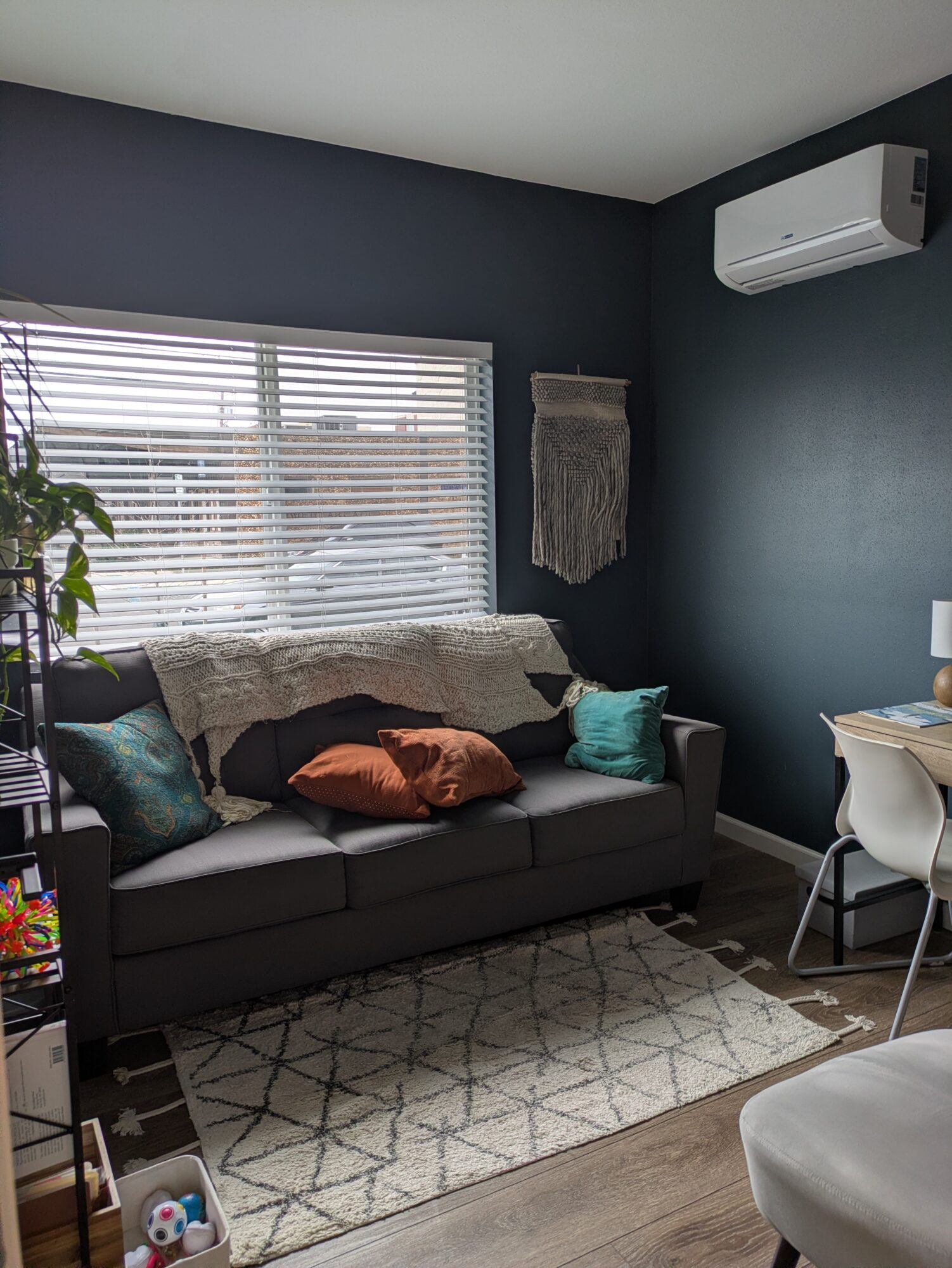 Living room with dark blue wall, window with blinds, gray sofa with pillows, wall hanging, air conditioner, and a small desk.