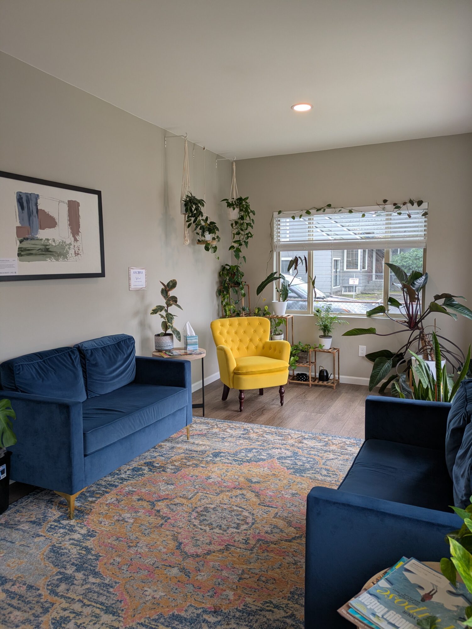 Living room with blue sofa, yellow armchair, large window with blinds, and various potted plants, rug, and wall art.