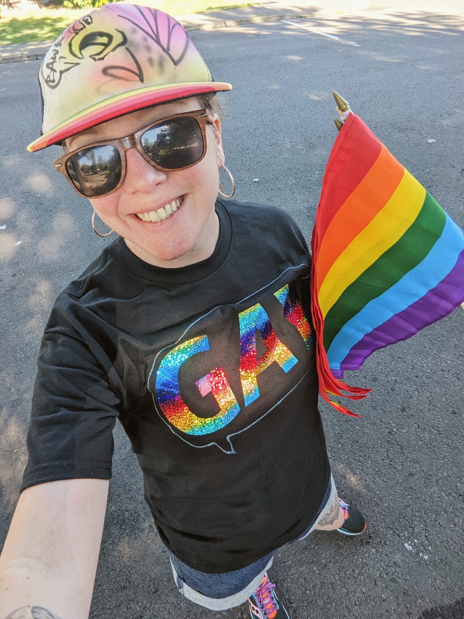 Person smiling, wearing sunglasses and a colorful hat, holding a rainbow pride flag, standing outdoors on a paved surface.