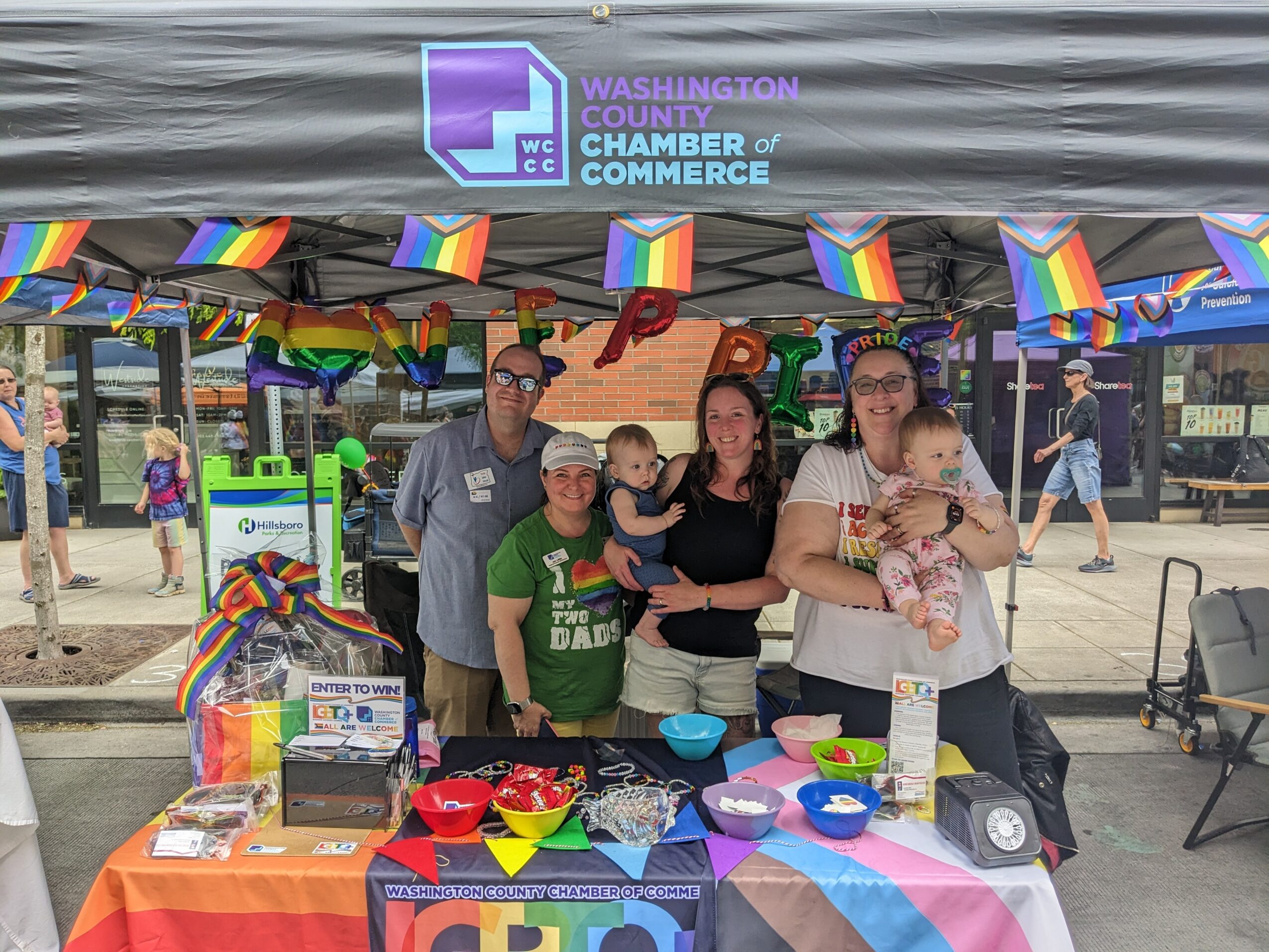 Group of people under a tent with rainbow flags, celebrating at an outdoor event, with a table of colorful items and a banner.