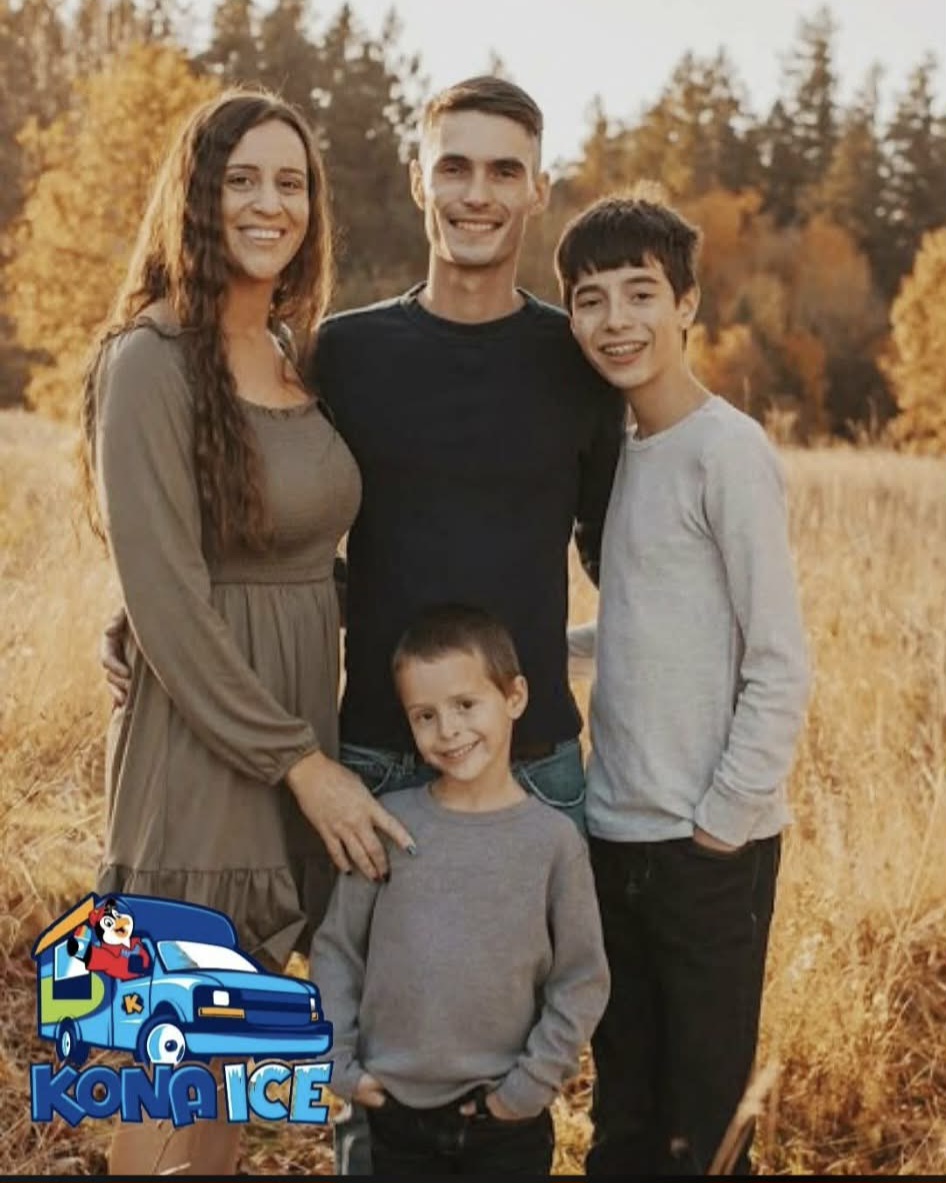 Family of four standing outdoors in a field with trees in the background, smiling, during autumn.