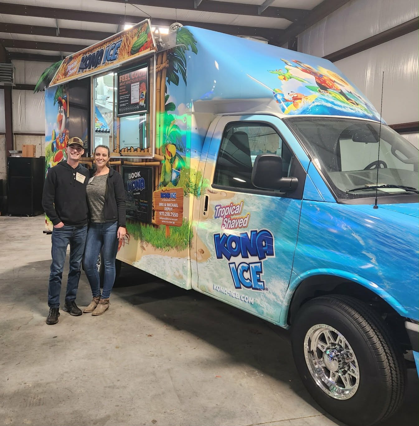 Two people stand in front of a colorful ice cream truck inside a warehouse, smiling at the camera.