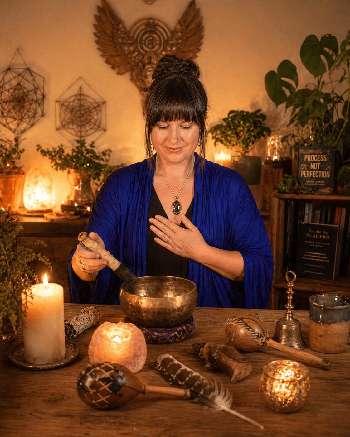 Woman with dark hair in a bun, wearing a blue shawl, holds a singing bowl with her right hand and places her left hand on her chest, seated at a table with candles and objects, in a warmly lit room with plants and decorative items.