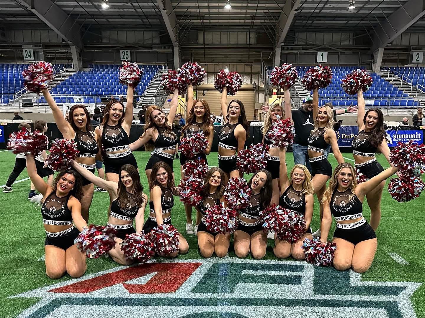 Group of cheerleaders in black outfits holding pom-poms, posing on a sports field inside a stadium.