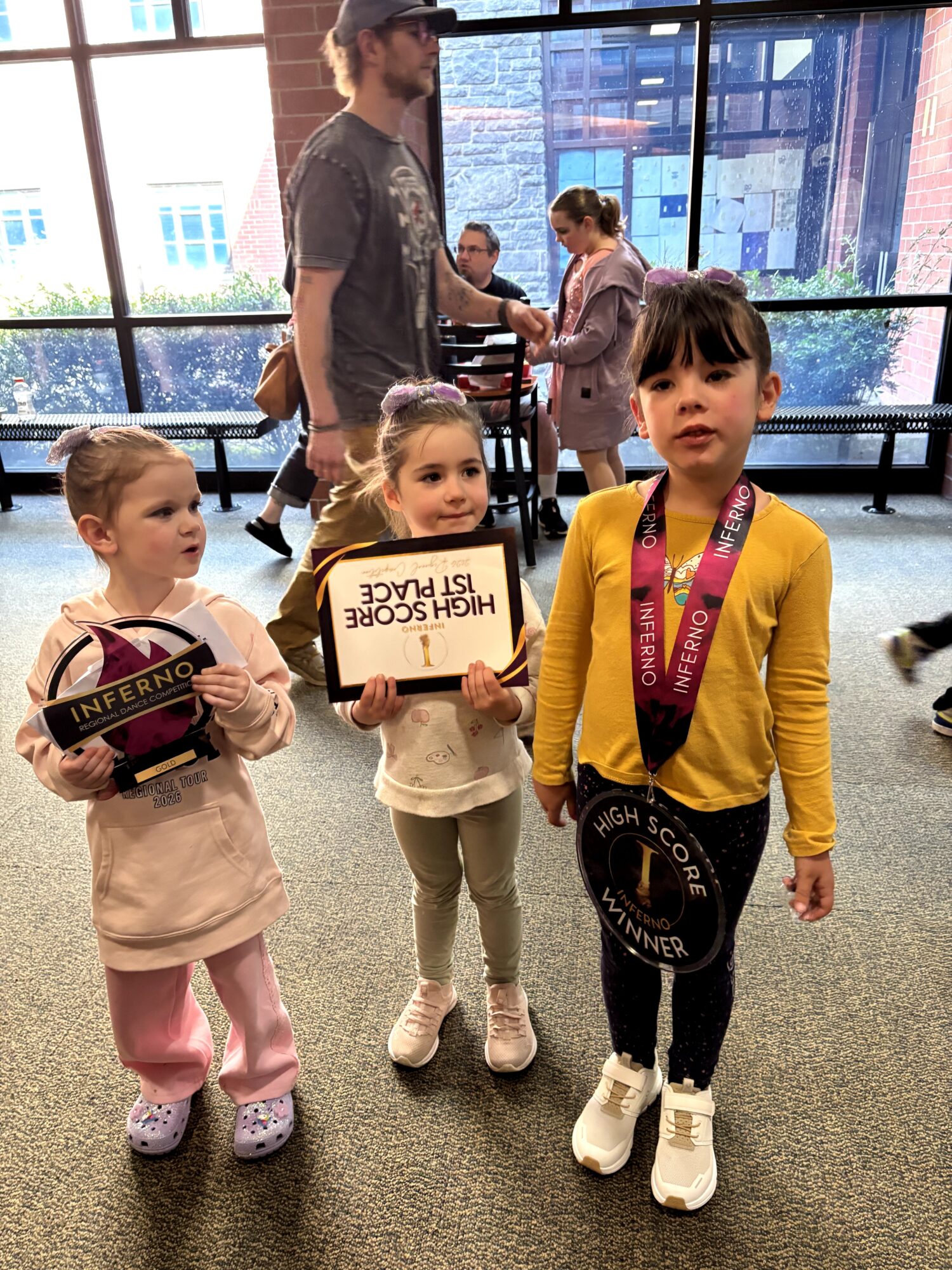 Three young girls standing indoors, one holding a sign, with adults and large windows in the background.