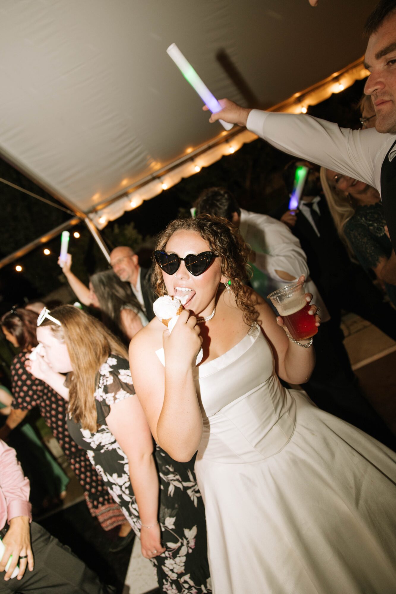 Woman in sunglasses and white dress holding a cupcake at a party, surrounded by people under a tent.