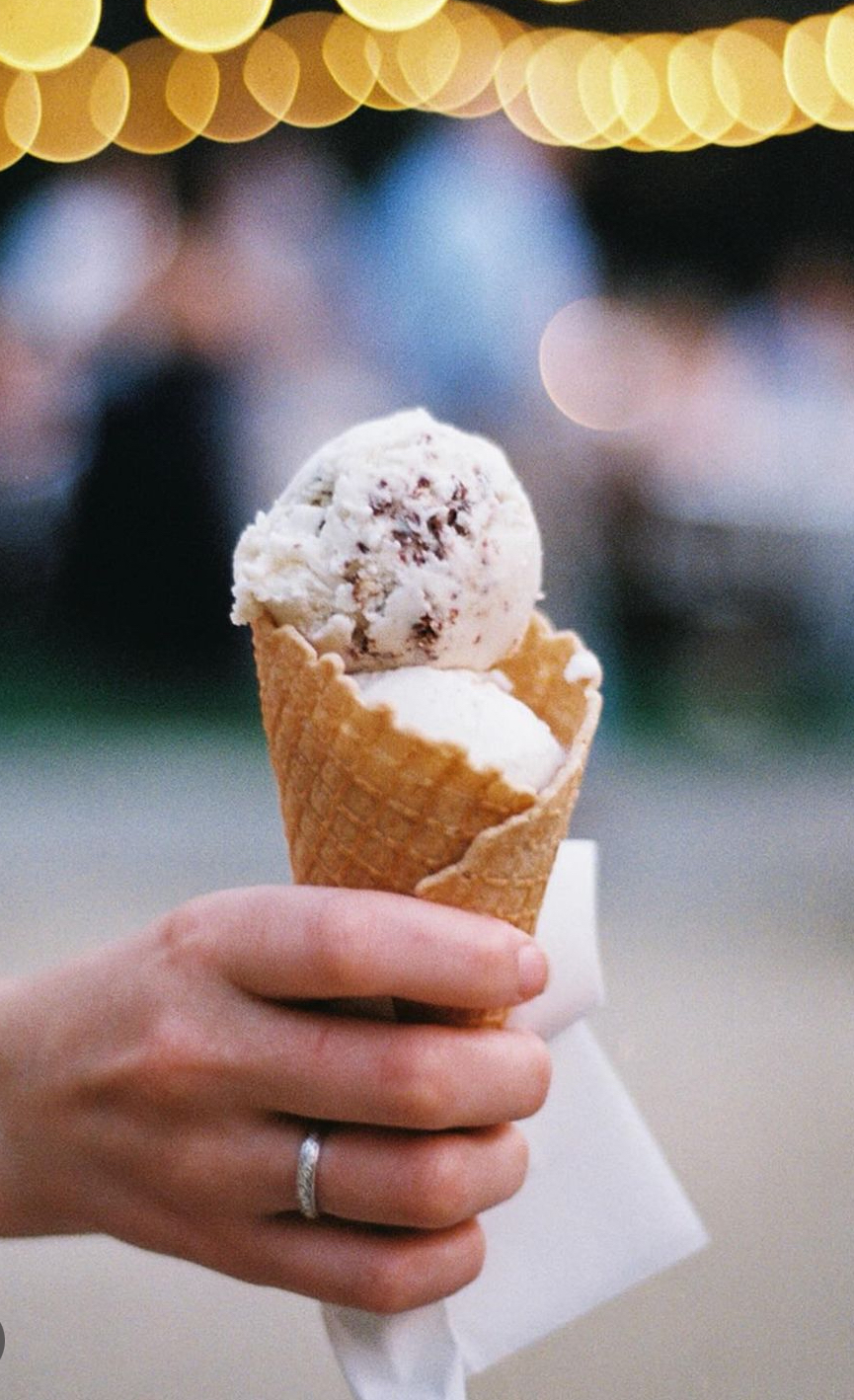 Hand holding an ice cream cone with two scoops, background with blurred lights and people.