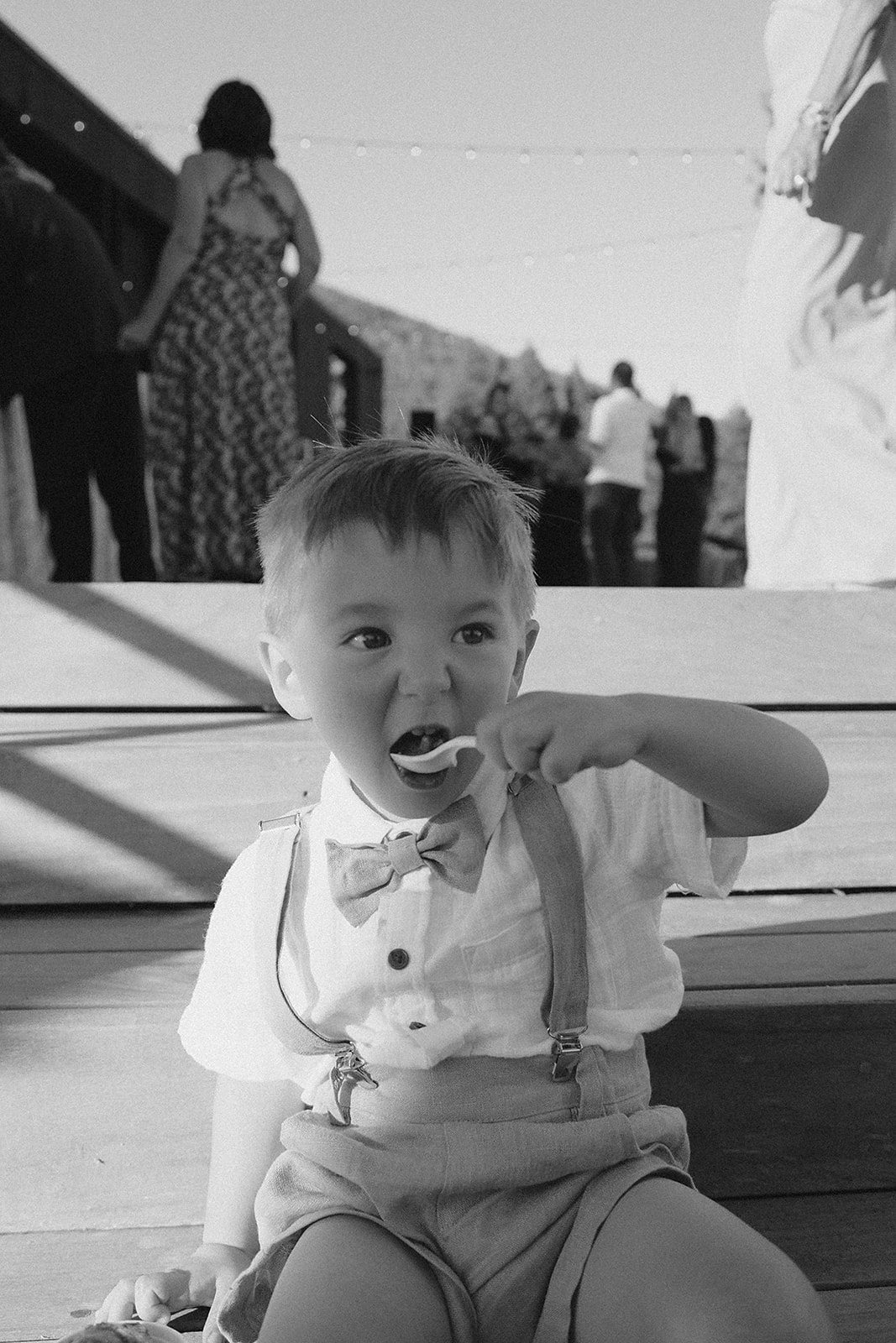 Young boy sitting on a bench, eating, with people and a building in the background, in black and white.